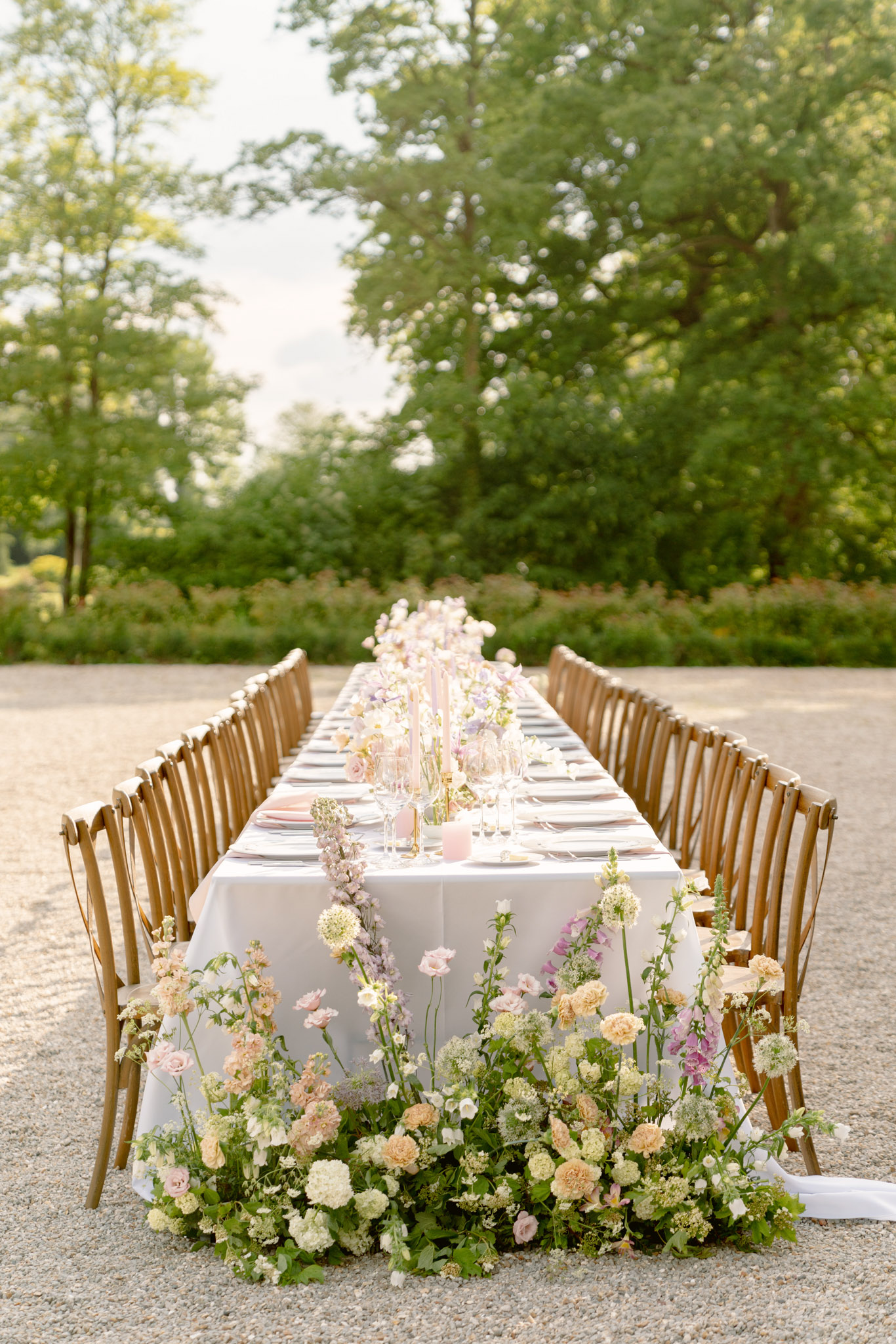 Long outdoor reception table with cascading blush roses, peach garden roses, lilac delphiniums, and pink taper candles