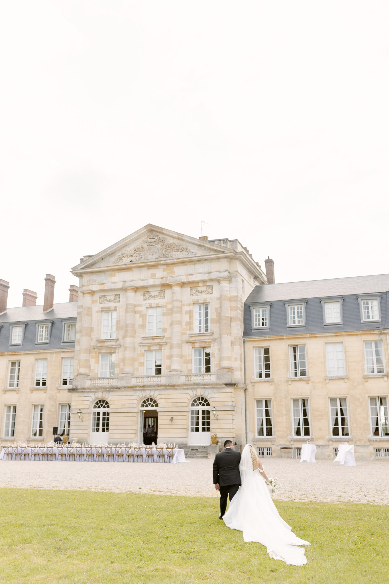 Couple walking toward chateau with reception tables bride in white gown with cathedral veil and bouquet