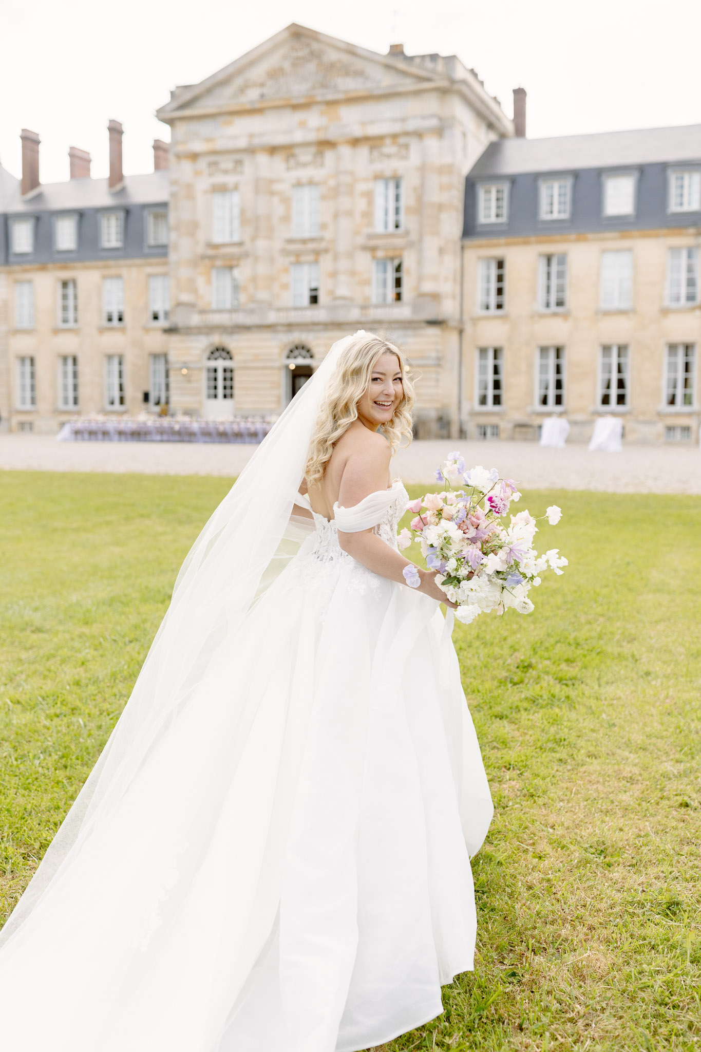 A bridal portrait taken outdoors on the lawn of a large French château, with the stone classical-style building visible in the background. The bride, a blonde woman with loose waves, is turned slightly away from the camera and glancing back over her shoulder with a smile, holding a loose, garden-style bouquet of white, blush pink, lavender, and lilac blooms including sweet peas and ranunculus. She wears an off-the-shoulder white ball gown with a fitted lace bodice and a full skirt, paired with a long cathedral-length veil that fans out behind her. In the background, rows of white ceremony chairs are arranged on the château forecourt. The shot is a three-quarter portrait with soft, even natural light.