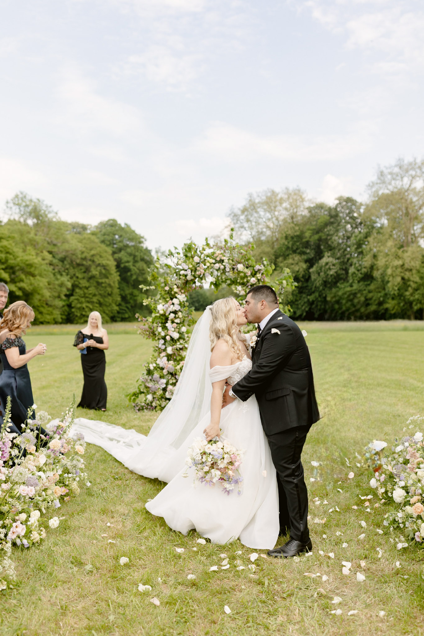 First kiss under circular floral arch with blush roses and lavender delphinium on garden lawn