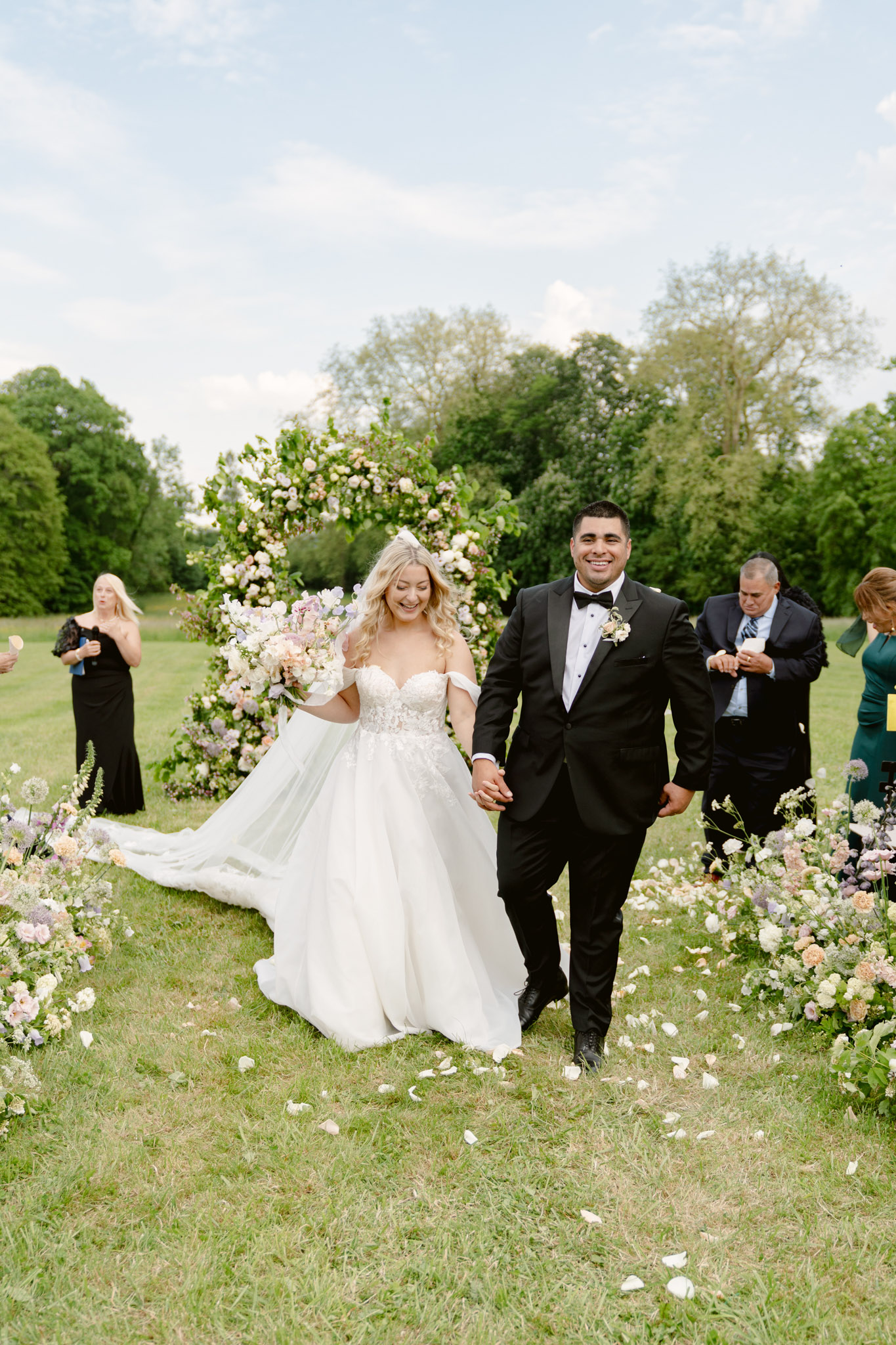 The newly married couple walks back up the aisle together following an outdoor ceremony on a grass lawn, holding hands and smiling. The bride wears an off-the-shoulder ballgown with a lace-embroidered bodice and a long tulle train, carrying a large loose bouquet in blush, lilac, and ivory tones including what appear to be garden roses, alliums, and soft trailing greenery. The groom wears a black tuxedo with a bow tie and a small ivory boutonnière. The aisle is lined with low garden-style floral arrangements in the same soft pastel palette of blush, lavender, ivory, and peach, with white flower petals scattered across the grass. Behind them stands a large circular floral arch densely covered in greenery and pastel blooms in blush and lilac. A small number of guests — including women in black and dark green gowns — are visible in the background near the arch. The overall floral and styling theme is romantic garden-style with a soft pastel palette. Medium-wide portrait shot.