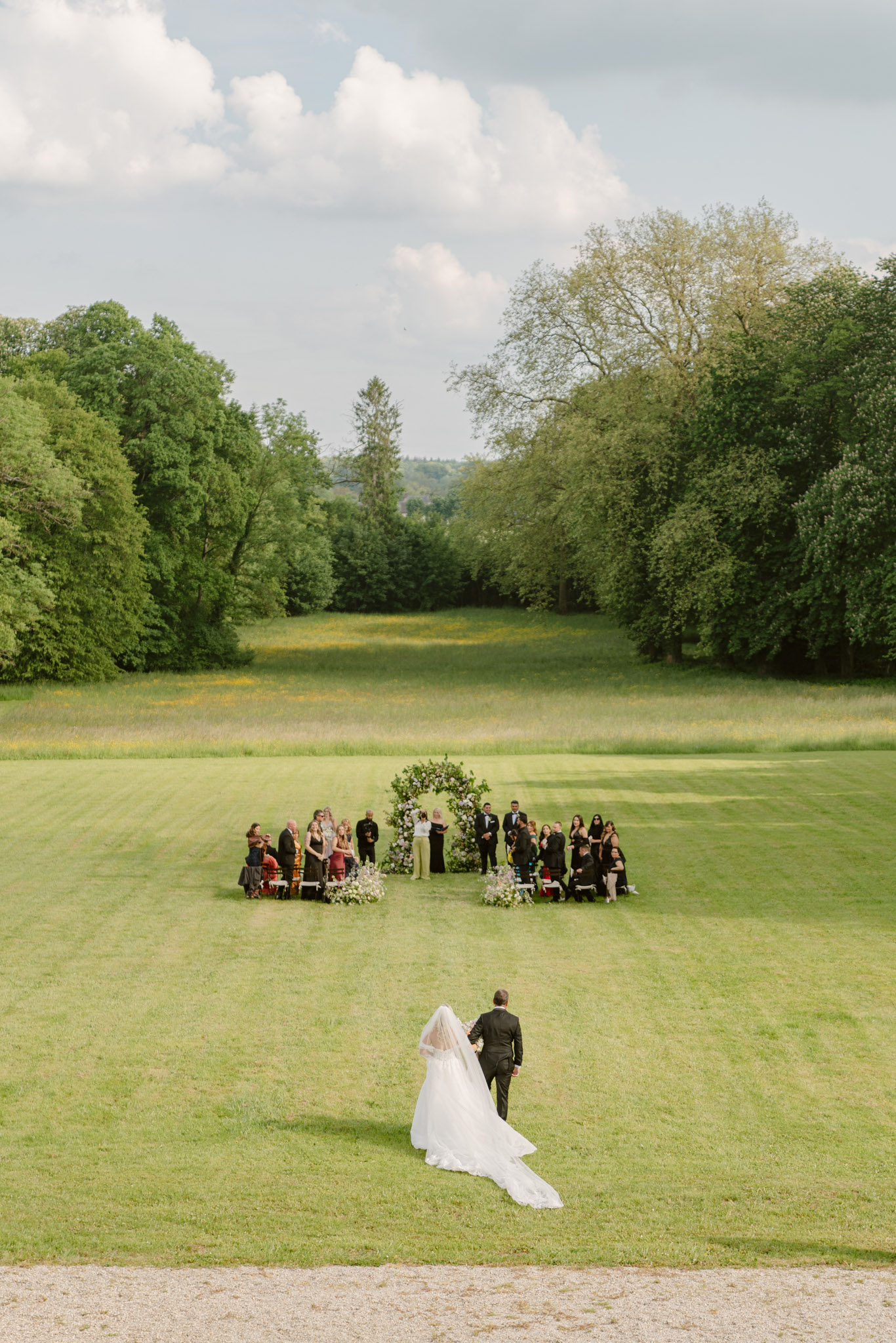 Elevated view of bride walking across lawn toward circular white and lavender floral arch with guests
