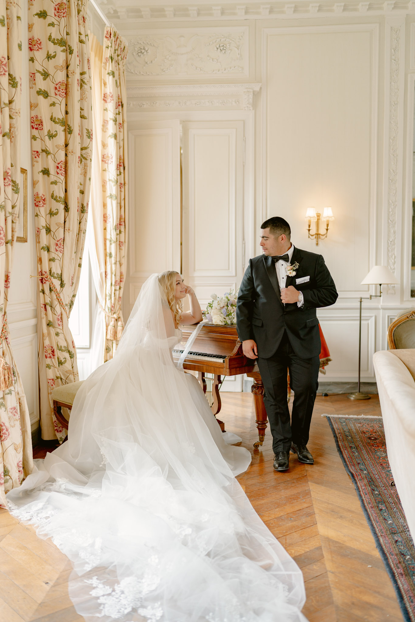 A couple portrait taken indoors in a classic French château salon, featuring the bride and groom together beside an antique wooden upright piano. The bride is seated on a bench leaning against the piano, wearing a full white ballgown with a long lace-trimmed cathedral veil that fans out dramatically across the herringbone parquet floor. She holds her hand to her cheek and looks up at the groom with a relaxed expression. The groom stands beside her in a black tuxedo with a bow tie and a white floral boutonnière. A loose bridal bouquet of soft white and pale lavender blooms rests on top of the piano. The room features white boiserie panelling with ornate plasterwork crown moulding, floor-length floral-print curtains in cream with red and green botanical motifs, a brass wall sconce, a traditional Persian-style area rug, and a gilded French sofa partially visible on the right. The styling is classic and formal. Medium wide shot capturing the full length of the bride's gown and veil.