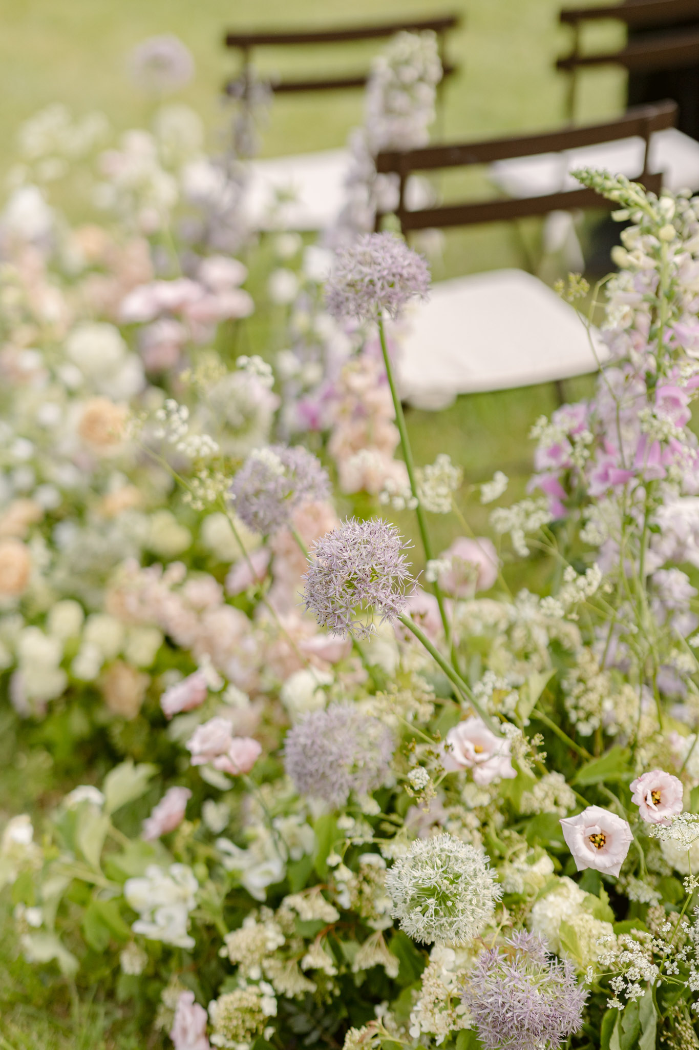 A close-up detail shot of an outdoor wedding ceremony aisle floral arrangement, with dark wooden folding chairs and white seat pads visible and softly blurred in the background. The arrangement features a wild, garden-style palette of lavender allium globe flowers, blush lisianthus, pale peach ranunculus, white Queen Anne's lace, pink foxglove, snapdragons, and trailing greenery. The overall color palette is soft and muted — lavender, blush, cream, and white — with an unstructured, meadow-inspired aesthetic. The shot is taken at a low angle, foregrounding the tallest flower stems against the open-air ceremony setting.