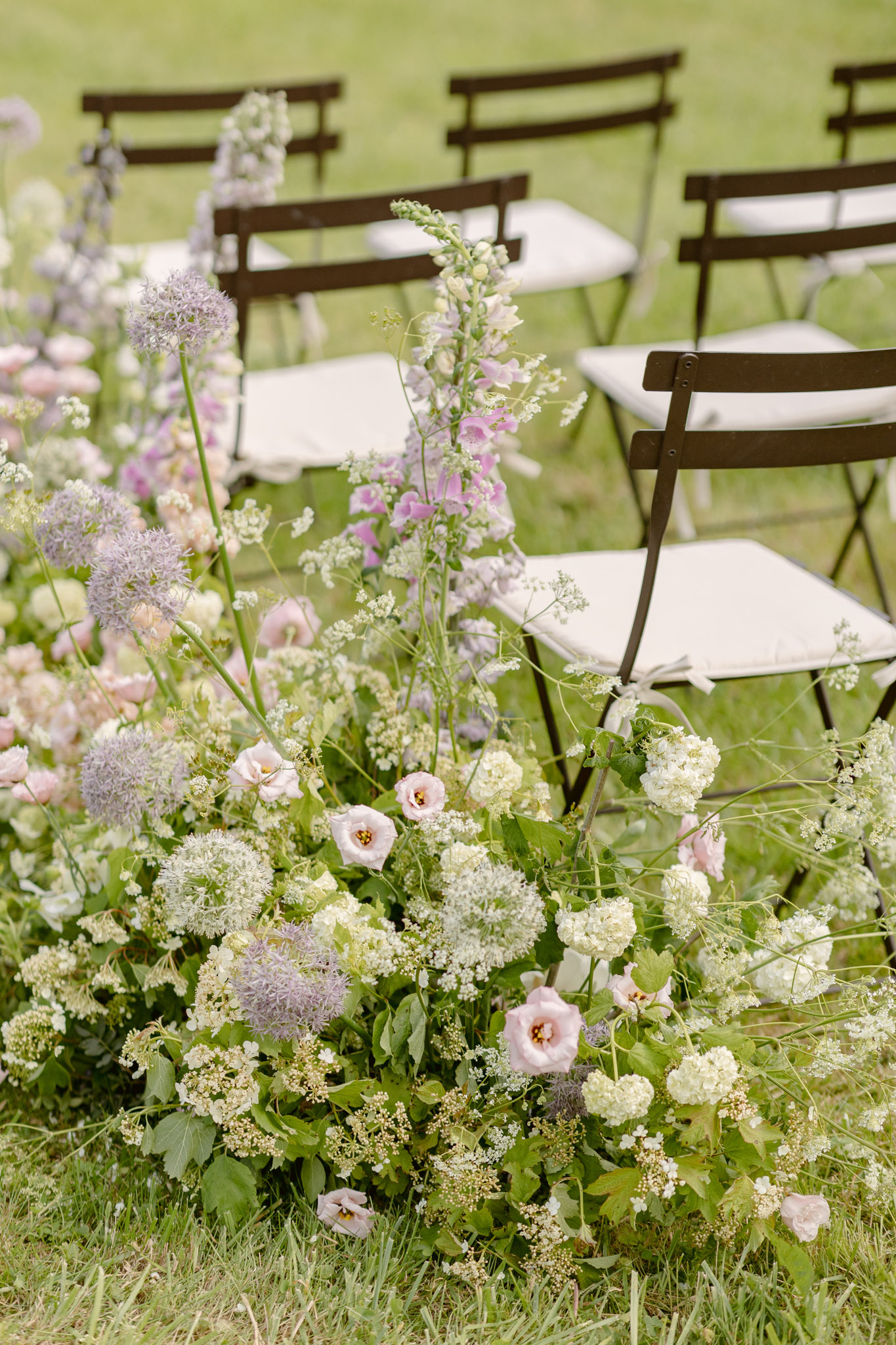 Ground-level ceremony aisle arrangement with blush lisianthus, purple allium, foxglove, and trailing greenery on lawn