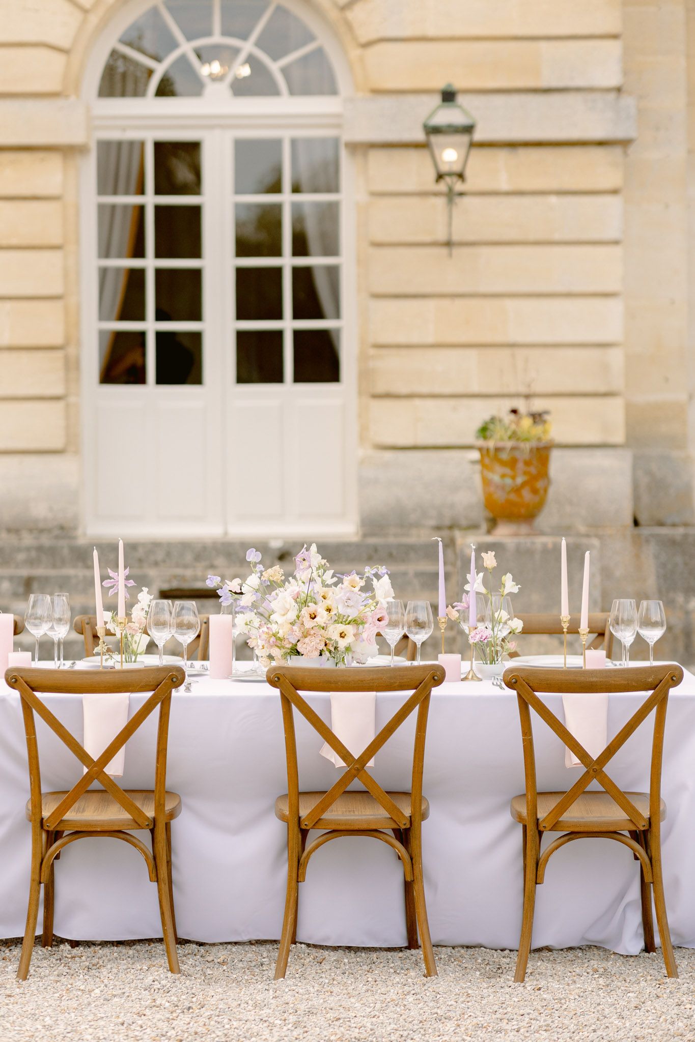 An outdoor reception table setup photographed in portrait orientation, positioned on a gravel courtyard directly in front of a classical French château facade with arched windows and a period wall lantern. The long table is covered with a white linen tablecloth and set with wine glasses in pairs, pale blush pink folded napkins, and gold taper candle holders holding slender lavender and blush pink candles. The central floral arrangement features cream garden roses, blush ranunculus, lavender sweet peas, and soft yellow blooms, with smaller individual stem bud vases scattered along the table. Natural wood cross-back chairs are positioned along one side of the table. The overall decor palette combines white, blush pink, and soft lavender with warm wood tones, following a classic French countryside aesthetic. The shot is taken from a slightly elevated angle looking straight at the table, functioning as a styled tablescape detail image. Potential venue feature image.