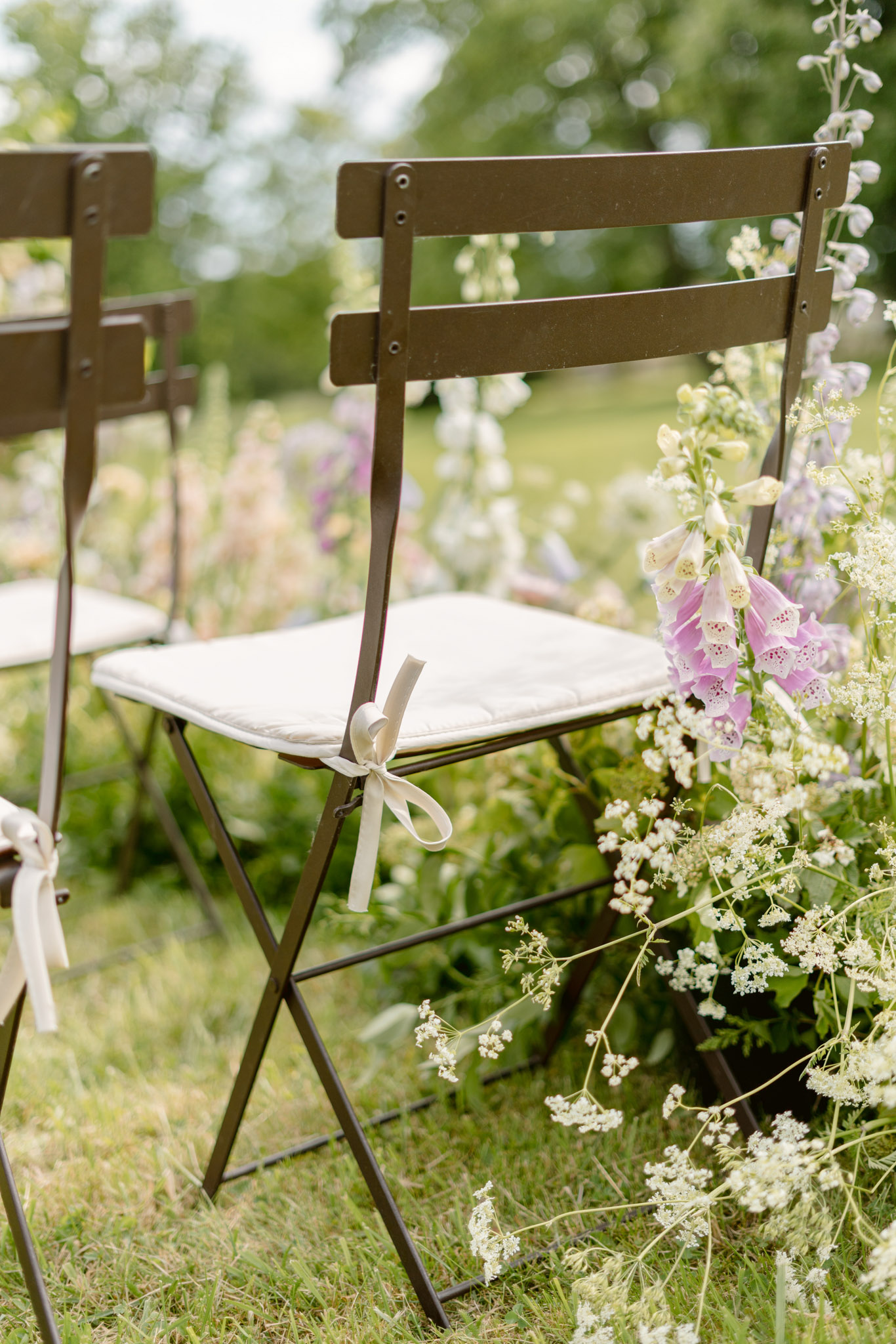 Wildflower aisle arrangement of pink foxgloves, white cow parsley, and purple delphiniums beside bistro chair