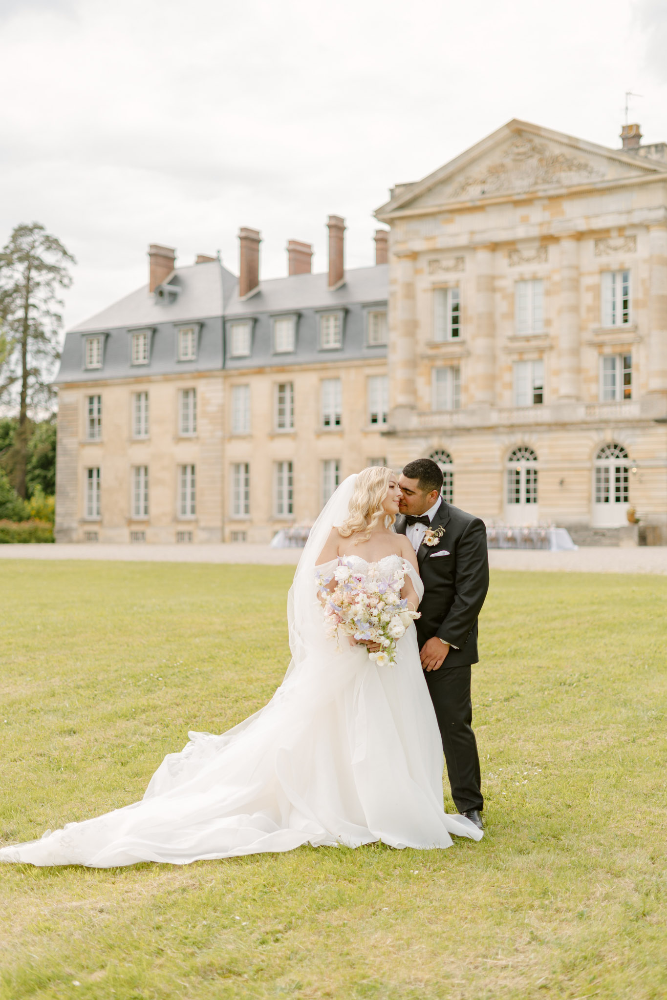 Groom kissing bride on cheek in front of limestone chateau with cathedral train spread on lawn