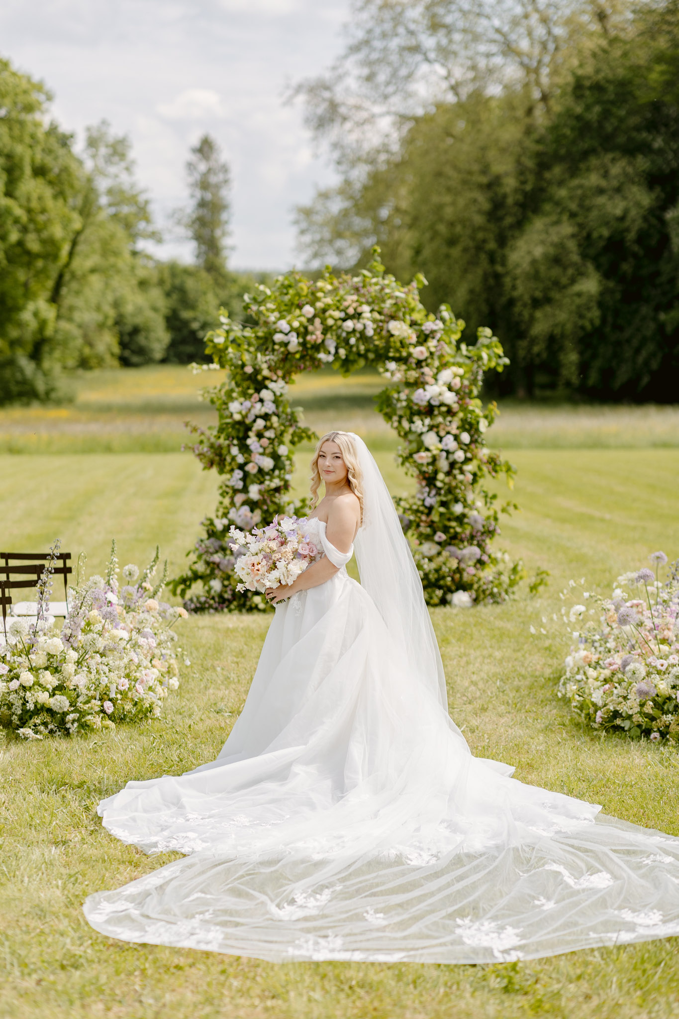 Bride in off-shoulder ballgown with cathedral train before circular arch of blush roses and lavender blooms