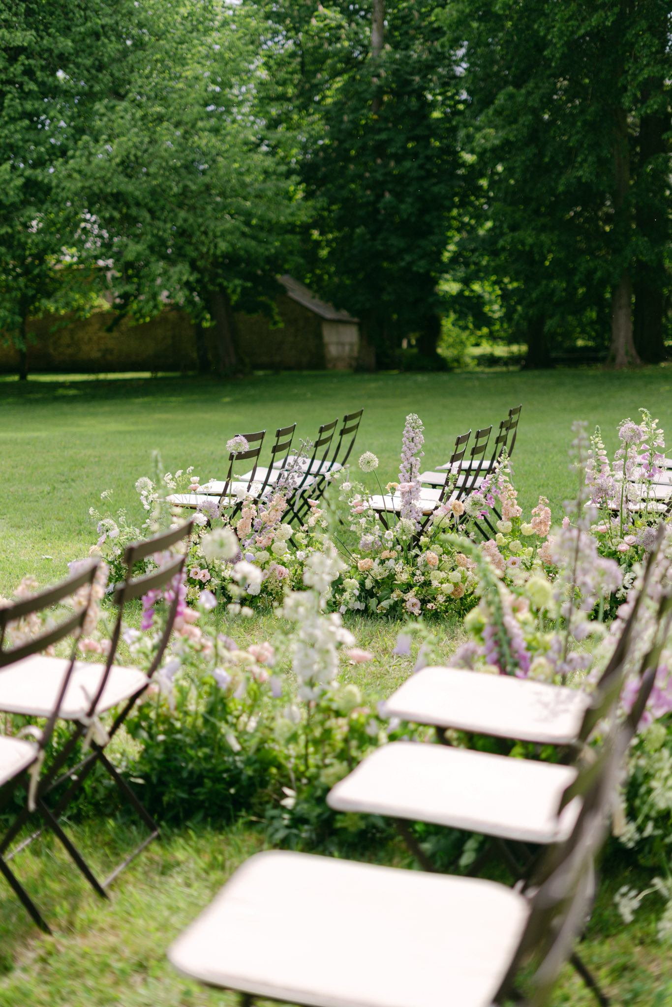 An outdoor wedding ceremony setup on a lawn, photographed from a low angle looking down the aisle toward the seating area. Dark metal folding chairs with cream or off-white seat cushions are arranged in rows on either side of a central aisle. The aisle is lined with abundant ground-level floral arrangements featuring blush garden roses, lavender and mauve delphiniums, white alliums, soft pink foxglove, ivory ranunculus, and trailing greenery, creating a lush garden-style border. The floral palette is a soft mix of blush, lilac, mauve, white, and pale green tones. The styling is romantic garden-inspired with a loose, naturalistic floral approach. A small stone outbuilding is partially visible in the background. Wide shot taken from ground level with shallow depth of field, keeping the foreground chairs slightly out of focus.