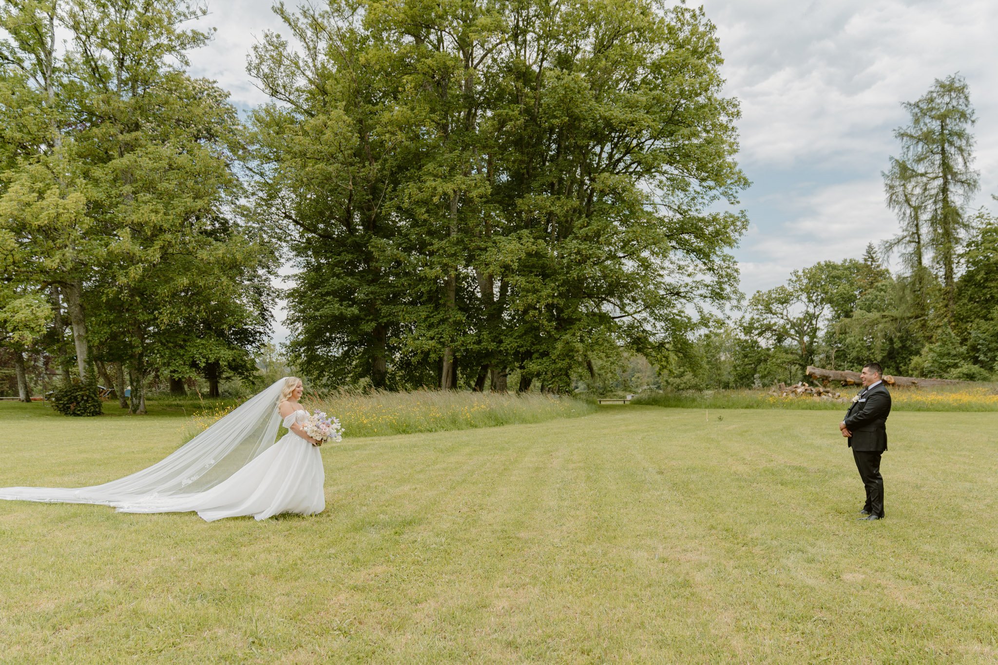 Bride approaches groom across wide lawn with cathedral veil trailing and pastel bouquet at chateau estate