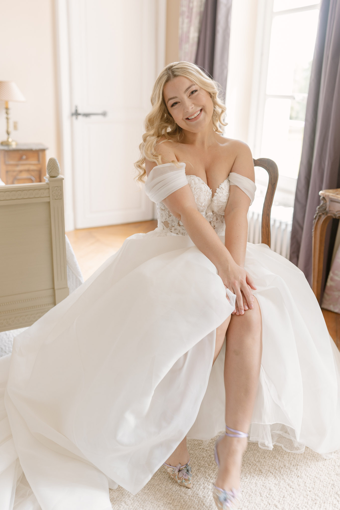 A getting-ready portrait of a bride seated on a carved wooden French-style chair in a bright, classically furnished room with warm white walls, parquet flooring, and grey-purple curtains framing a window. She is wearing an off-the-shoulder white ball gown with a lace appliqué bodice and a full organza skirt, and is in the process of fastening a pale blue satin garter on her thigh. Her footwear consists of embellished heels with pale blue ankle straps and decorative bow detailing, referencing the 'something blue' tradition. She has long blonde wavy hair worn loose and is smiling directly at the camera. The room features a grey-painted vintage dresser and a table lamp, suggesting a château or manor house bridal suite setting. The image is a medium portrait shot taken in soft natural window light with a warm, airy tone.