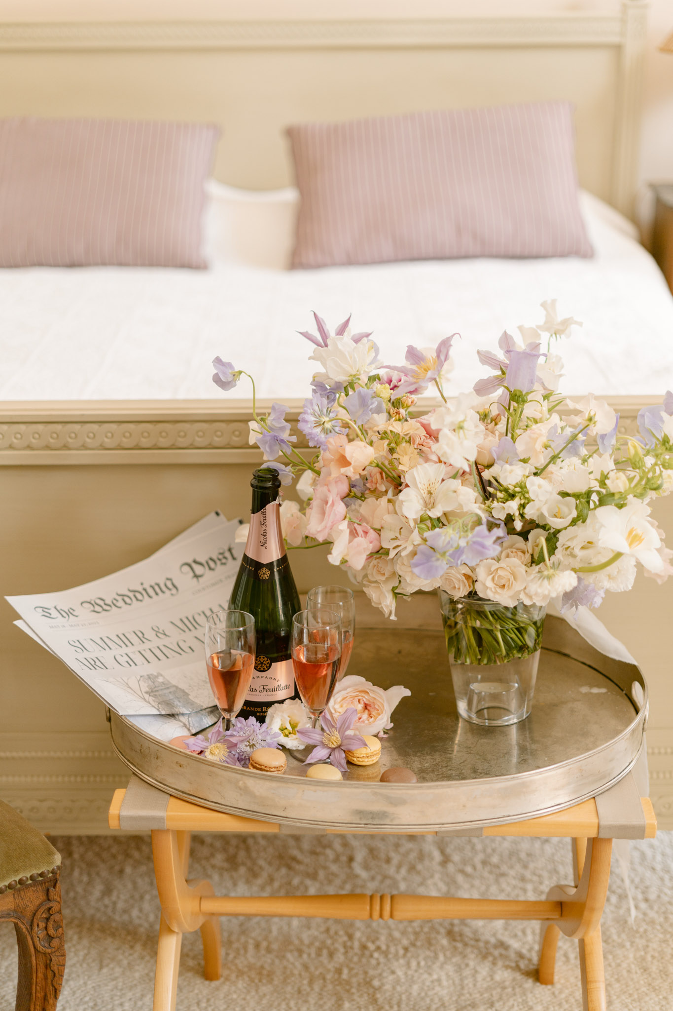 A styled flat-lay-style detail shot of a bridal suite getting-ready setup, photographed from a slightly elevated angle in a softly lit interior bedroom. A round silver tray rests on a wooden folding luggage rack and holds a bottle of Nicolas Feuillatte Grande Réserve rosé champagne, two filled champagne flutes with pink bubbling liquid, a custom personalised newspaper titled 'The Wedding Post' with the couple's names 'Summer & Michael' visible, several French macarons in pastel shades, and scattered loose clematis and garden rose blooms. A large arrangement of flowers sits beside the tray in a clear glass vase, featuring ivory garden roses, blush sweet peas, lavender-purple clematis, and white lisianthus. The bed in the background is dressed in white linens with mauve-pink striped pillowcases, and the room has cream-painted panelled walls and furniture in a classic French interior style. The overall decor palette is blush, ivory, and lavender, consistent with a soft romantic styling theme.