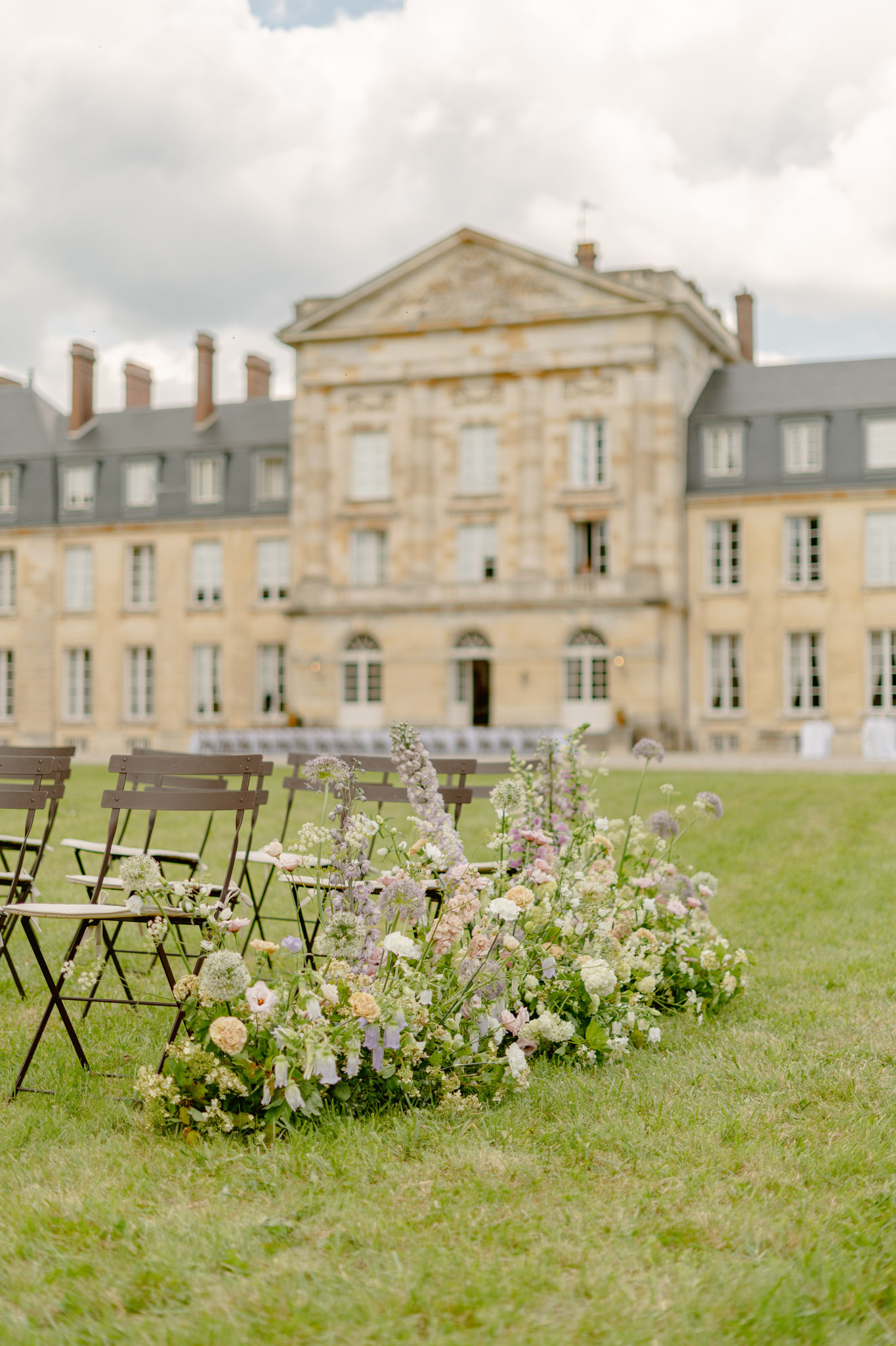 An outdoor wedding ceremony setup on the grounds of a large French château, with rows of dark brown folding chairs arranged on an open lawn facing the building. In the foreground, a ground-level floral arrangement serves as an aisle marker, composed of peach garden roses, lavender and lilac delphiniums, white queen anne's lace, mauve lisianthus, allium, and soft green foliage in a loose, organic style. The floral palette is soft and muted — blush, ivory, lavender, and peach. No people are present; the scene appears to be set up prior to the ceremony. The shot is taken from a low angle close to the floral arrangement, with the château softly blurred in the background showing a classical stone facade with mansard roofs and multiple wings. Potential venue feature image.