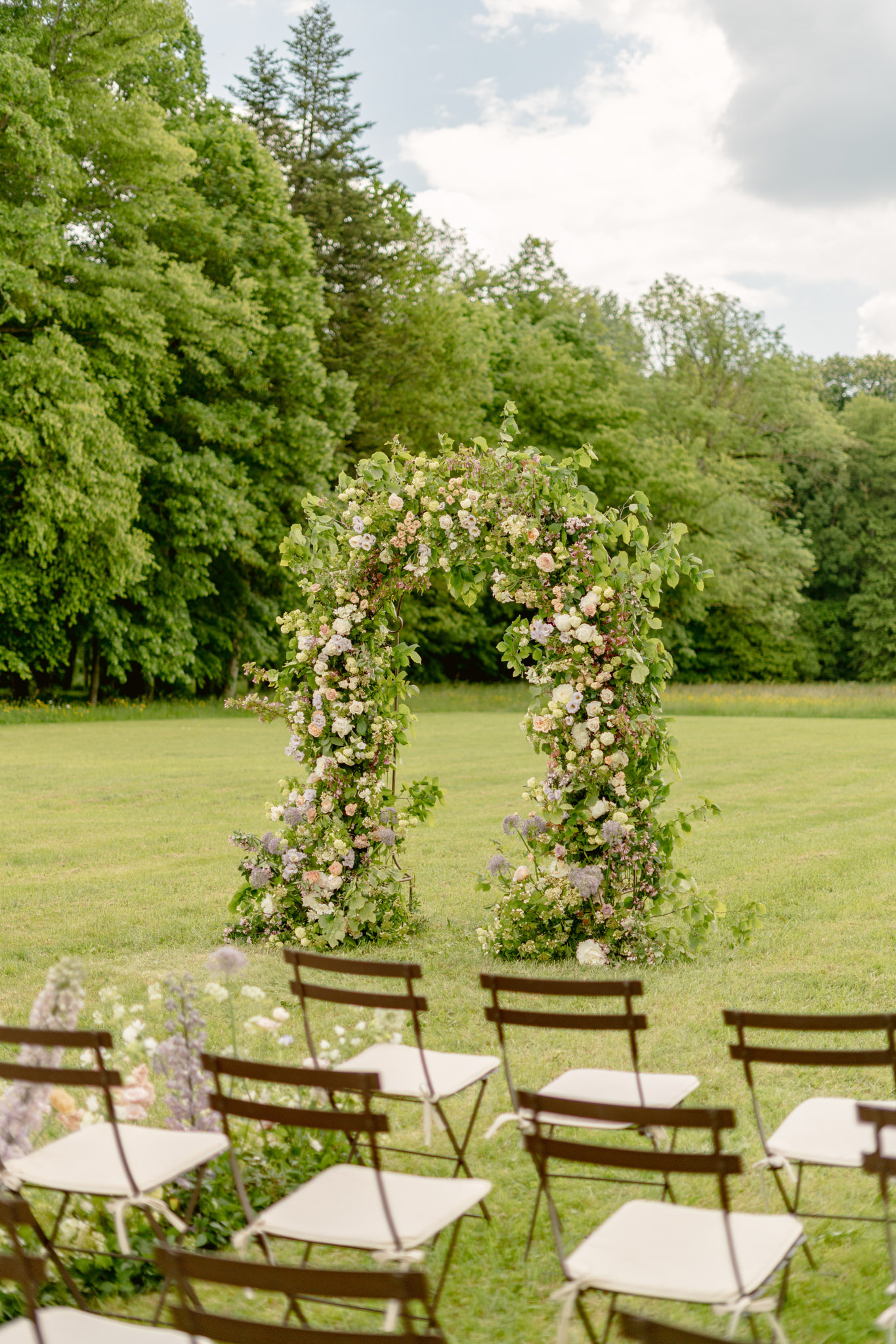 Organic floral arch of blush roses, mauve alliums, and lavender anemones with bistro chairs on lawn