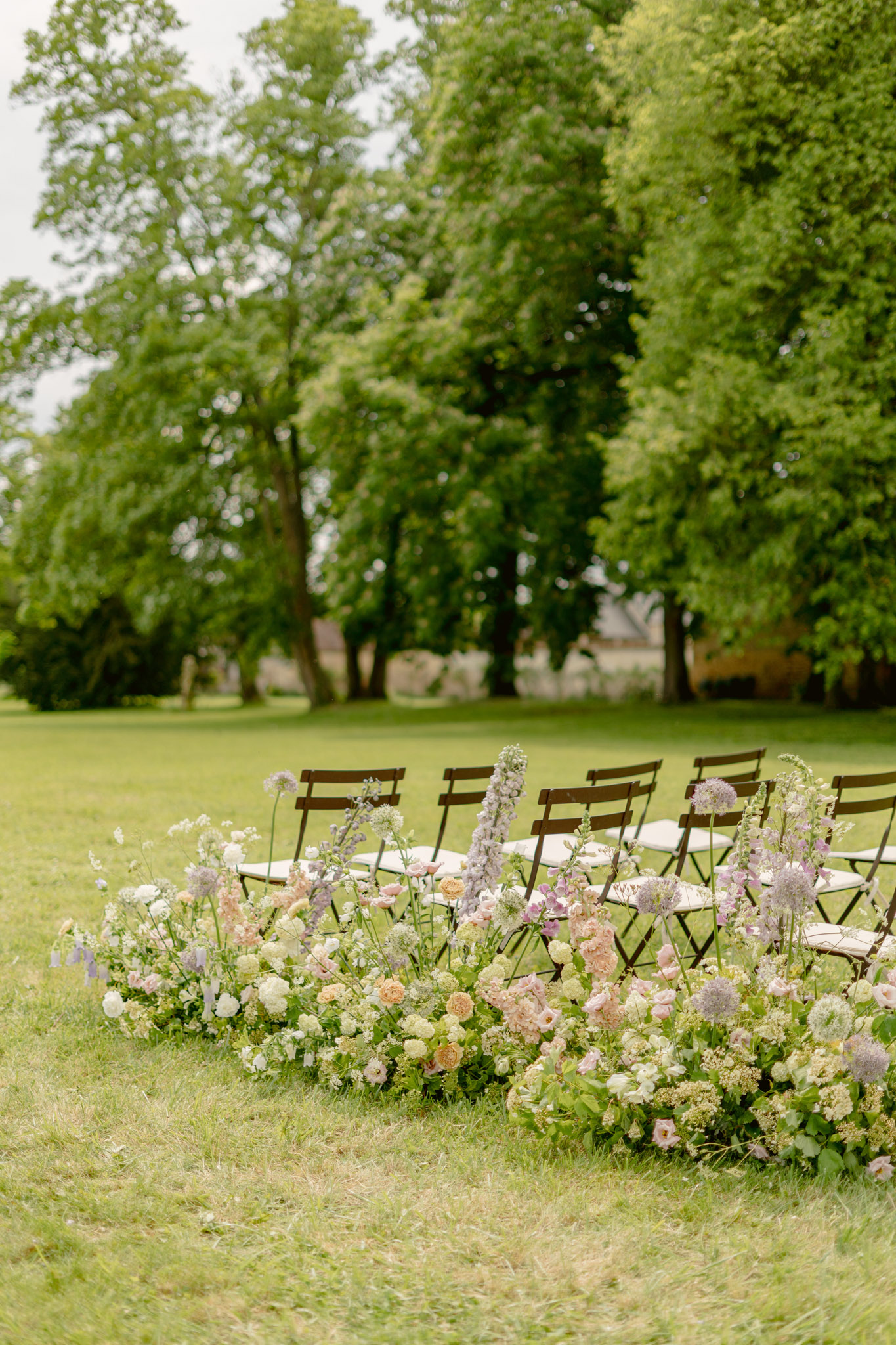 Garden-style aisle flowers of blush roses, lilac delphiniums, and alliums with brown bistro chairs on lawn