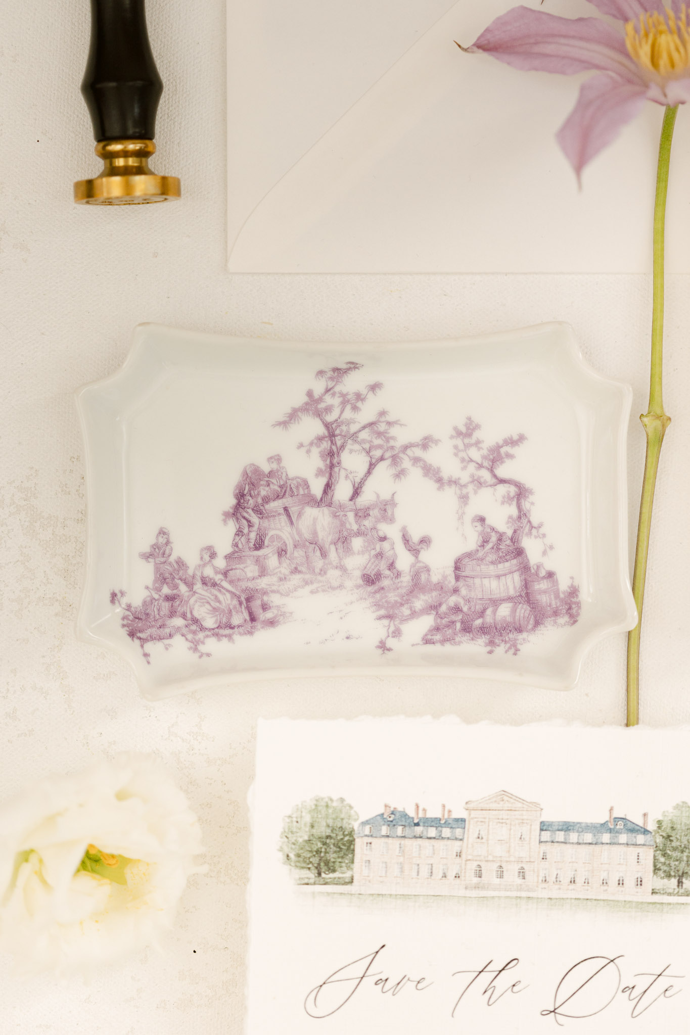 A flat lay detail shot of wedding stationery and decorative objects arranged on a white textured surface. The centerpiece is a white porcelain tray featuring a mauve-purple toile de Jouy pastoral scene. Below it, a deckle-edged save the date card is partially visible, printed with a watercolor illustration of a French château and copper-toned calligraphy reading 'Save the Date.' A black and gold wax seal stamp appears in the upper left corner, a cream peony bloom is visible in the lower left, and a single mauve clematis flower with its stem extends along the right side. The overall color palette is soft ivory, mauve-purple, and copper, with a classic French-inspired aesthetic.