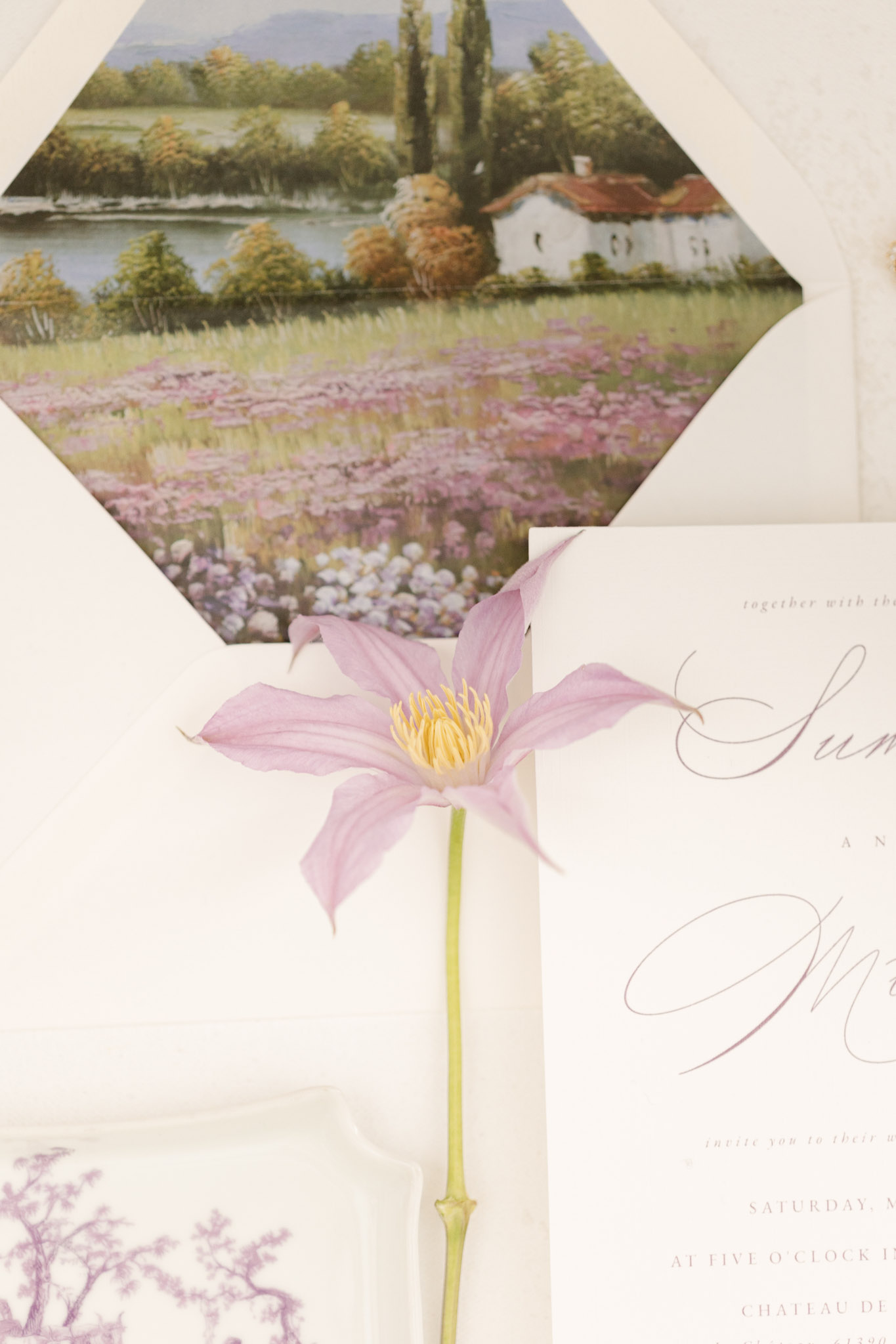Wedding stationery flat lay with calligraphy invitation, painted landscape envelope liner, and mauve clematis bloom