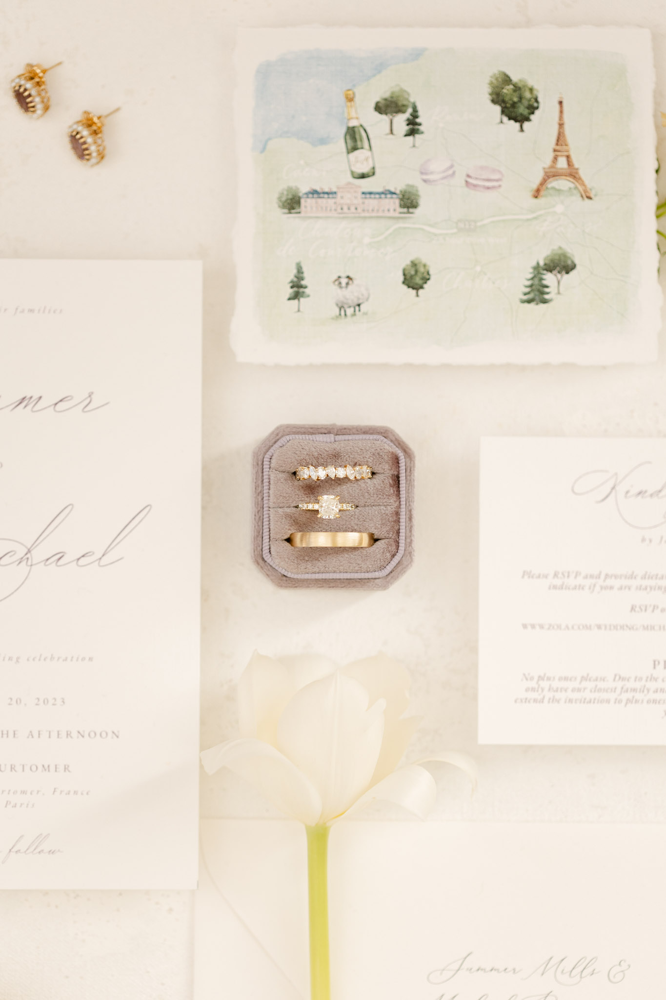 Stationery flat lay with watercolor venue map diamond rings in grey velvet box gold earrings and white tulip on marble