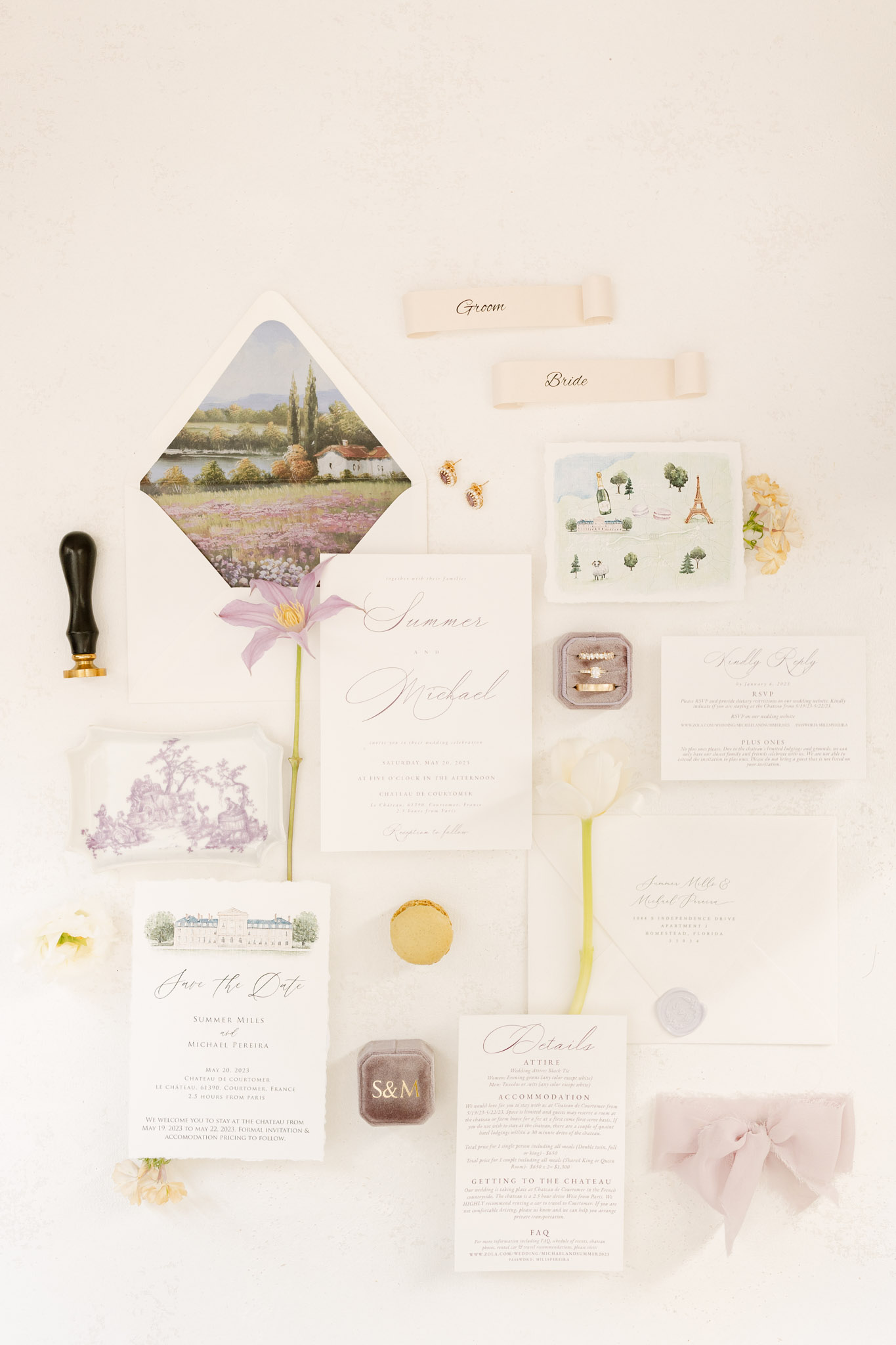 A flat lay detail shot of a complete wedding stationery suite for Summer Mills and Michael Pereira, set against a cream background. The suite includes a main invitation card in white with calligraphy script, a save-the-date card featuring a watercolor illustration of a French château, an envelope with a painted lavender field landscape liner, an RSVP card, a details card, and an addressed outer envelope. Additional styling elements include a dusty mauve velvet ring box monogrammed with 'S&M' containing wedding rings, a second velvet ring box with pearl and gold stacking rings, small gold bee stud earrings, a gold macaron, a wax seal stamp with black and gold handle, a pale lavender wax seal, a watercolor illustration card featuring the Eiffel Tower and French landmarks, a purple toile de Jouy printed notecard, blush silk ribbon curls labeled 'Bride' and 'Groom,' a single pale purple clematis flower stem, small cream and yellow florals, and a dusty pink bow. The overall palette is soft blush, lavender, sage green, and gold, with a classic French-inspired design aesthetic.