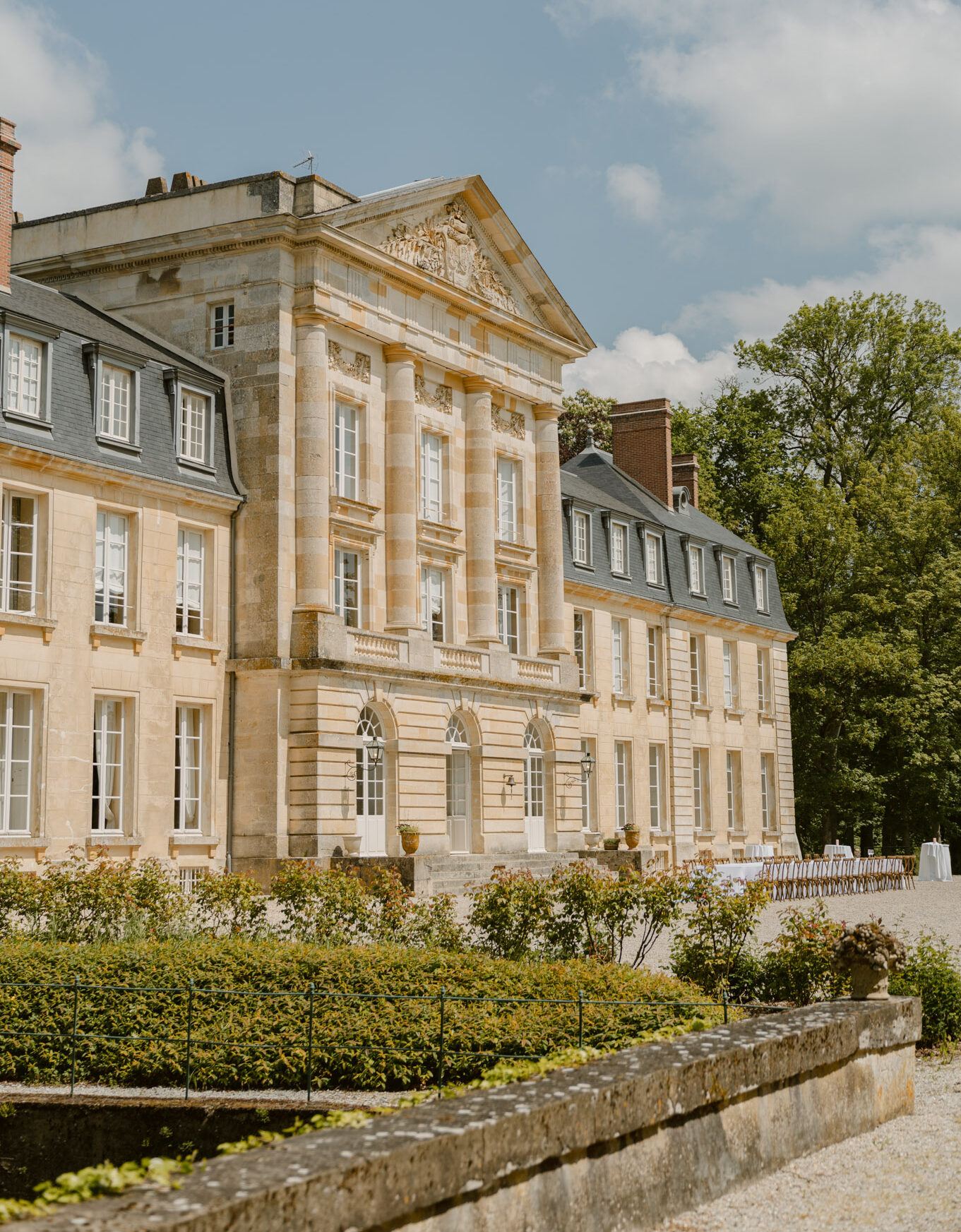 Wide exterior shot of a French classical château with warm sandstone façade, slate mansard roofs, and a central pediment featuring decorative carved relief work above pilastered columns. The building spans multiple wings across the frame. To the right of the main entrance, rows of wooden chairs are arranged in a ceremony setup on a gravel courtyard, with what appears to be a white-clothed cocktail or welcome table nearby, indicating the space is dressed for a wedding ceremony outdoors. Trimmed hedgerows and a stone-edged reflecting pool or parterre border the foreground. The overall style of the event setup visible is classic and understated, with natural wood chairs against the formal French architecture. Potential venue feature image.