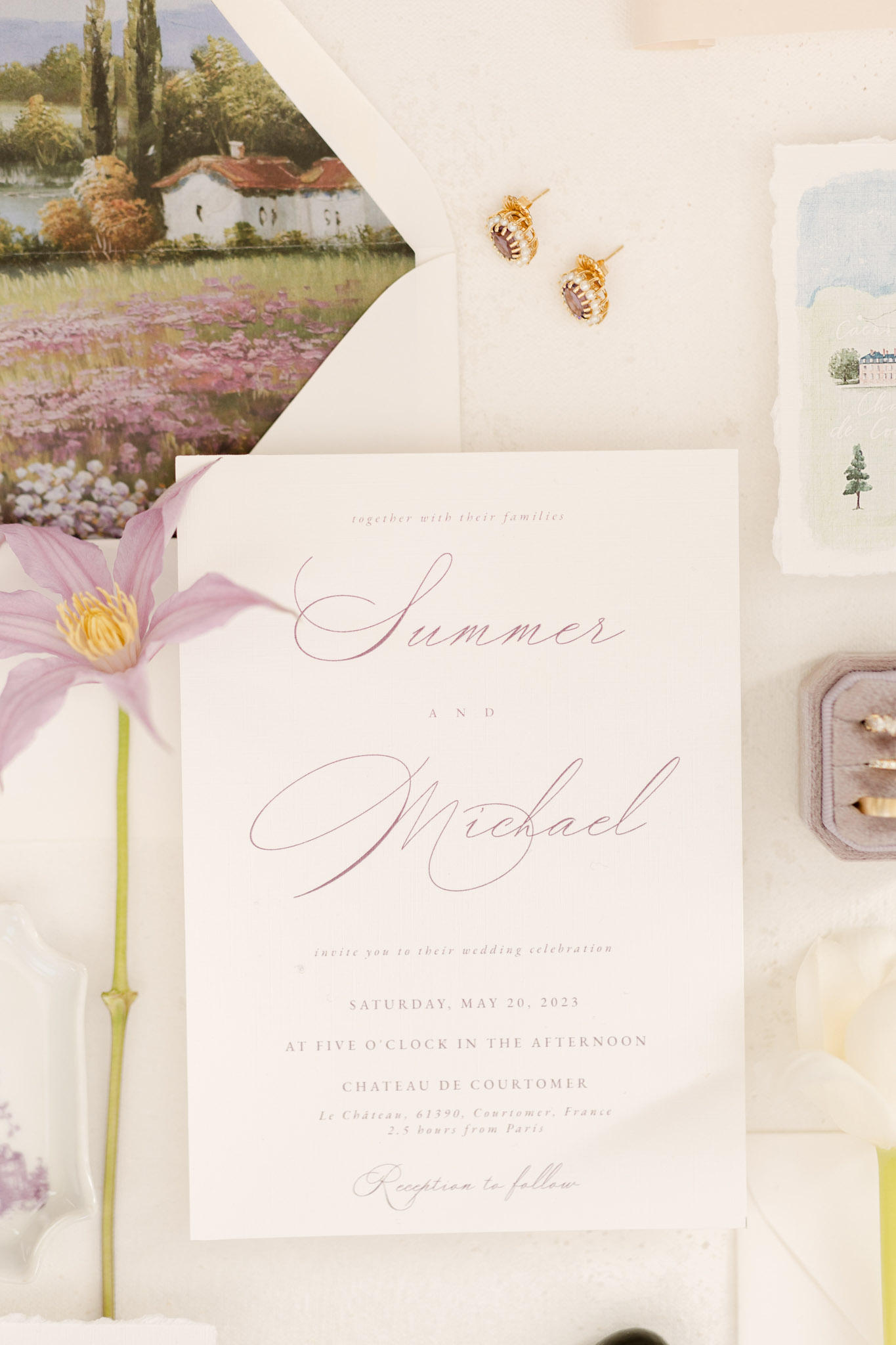 A flat lay detail shot of a wedding stationery suite for Summer and Michael, dated Saturday, May 20, 2023, at Chateau de Courtomer, Le Château, 61390, Courtomer, France. The central invitation card features an ivory background with dusty rose calligraphy script for the names and serif print for the event details. Surrounding items include an envelope liner printed with a painterly landscape illustration of a lavender field and countryside scene, a watercolor venue illustration card with deckled edges, a grey velvet ring box holding wedding bands, a pair of small gold stud earrings with purple gemstones, and a single pale lavender clematis bloom placed along the left side of the arrangement. The overall palette is soft ivory, dusty rose, lilac, and grey, consistent with a classic, romantic French wedding aesthetic.