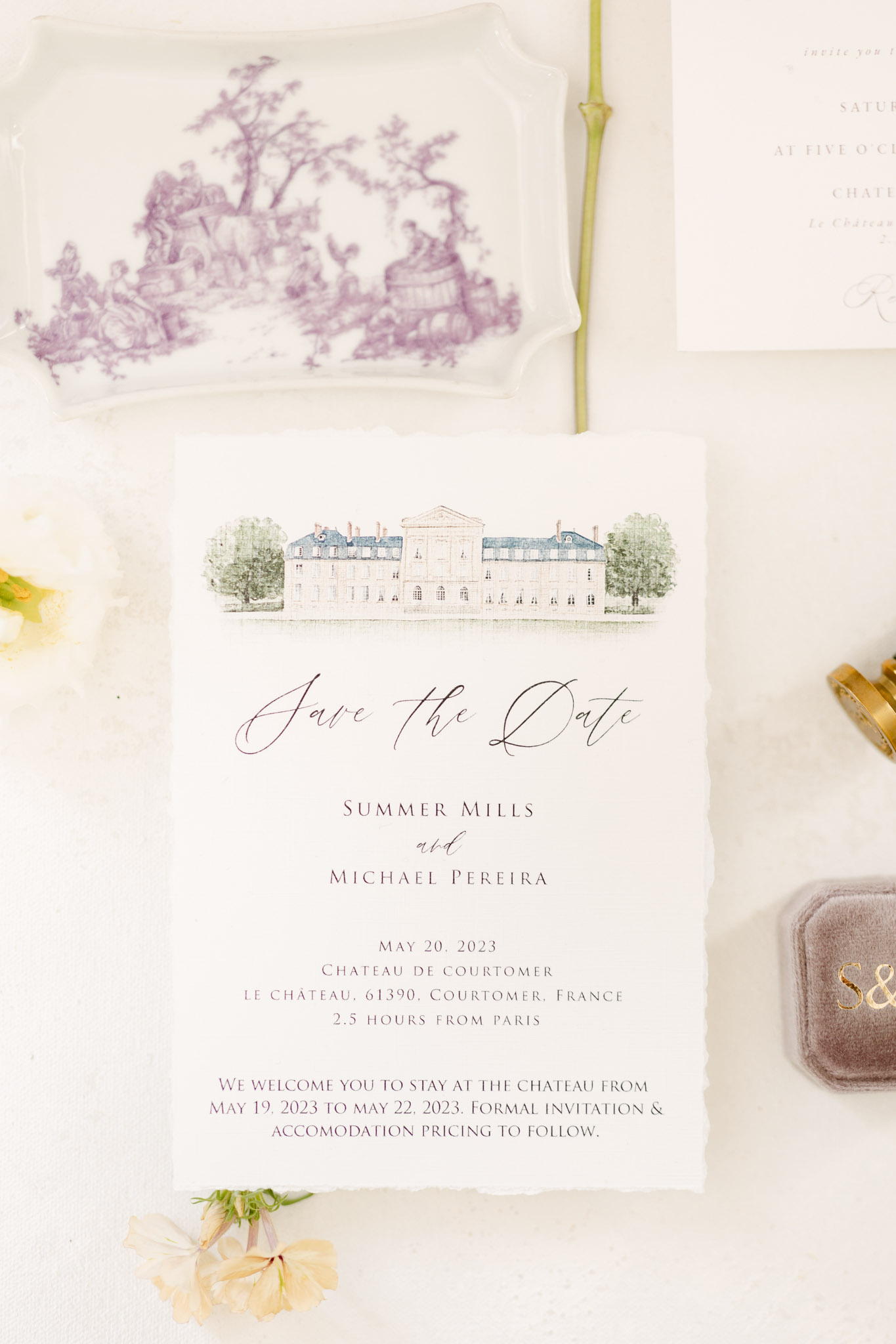 A flat lay of a wedding stationery suite styled on a white surface. The central piece is a save-the-date card for Summer Mills and Michael Pereira, dated May 20, 2023, at Château de Courtomer, Courtomer, France, featuring a watercolor illustration of the château facade at the top and script lettering in deep burgundy and dark ink. Surrounding the card are a white ceramic tray with a purple toile de Jouy pattern, a partial formal invitation card in ivory, a gold ring box, a taupe velvet ring box monogrammed with 'S&' in gold, and small loose peach and cream florals scattered at the edges. The overall palette is soft white, burgundy, gold, and dusty purple, reflecting a classic French château wedding aesthetic.