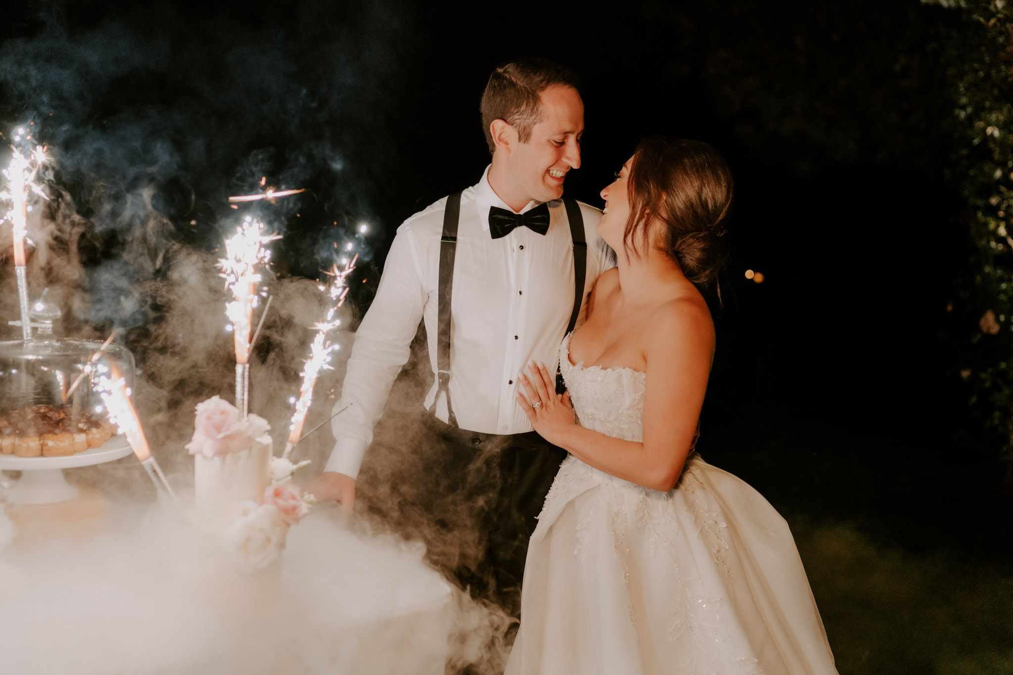 Couple laughing at sparkler-lit cake cutting with blush rose two-tier cake and dry ice fog at night