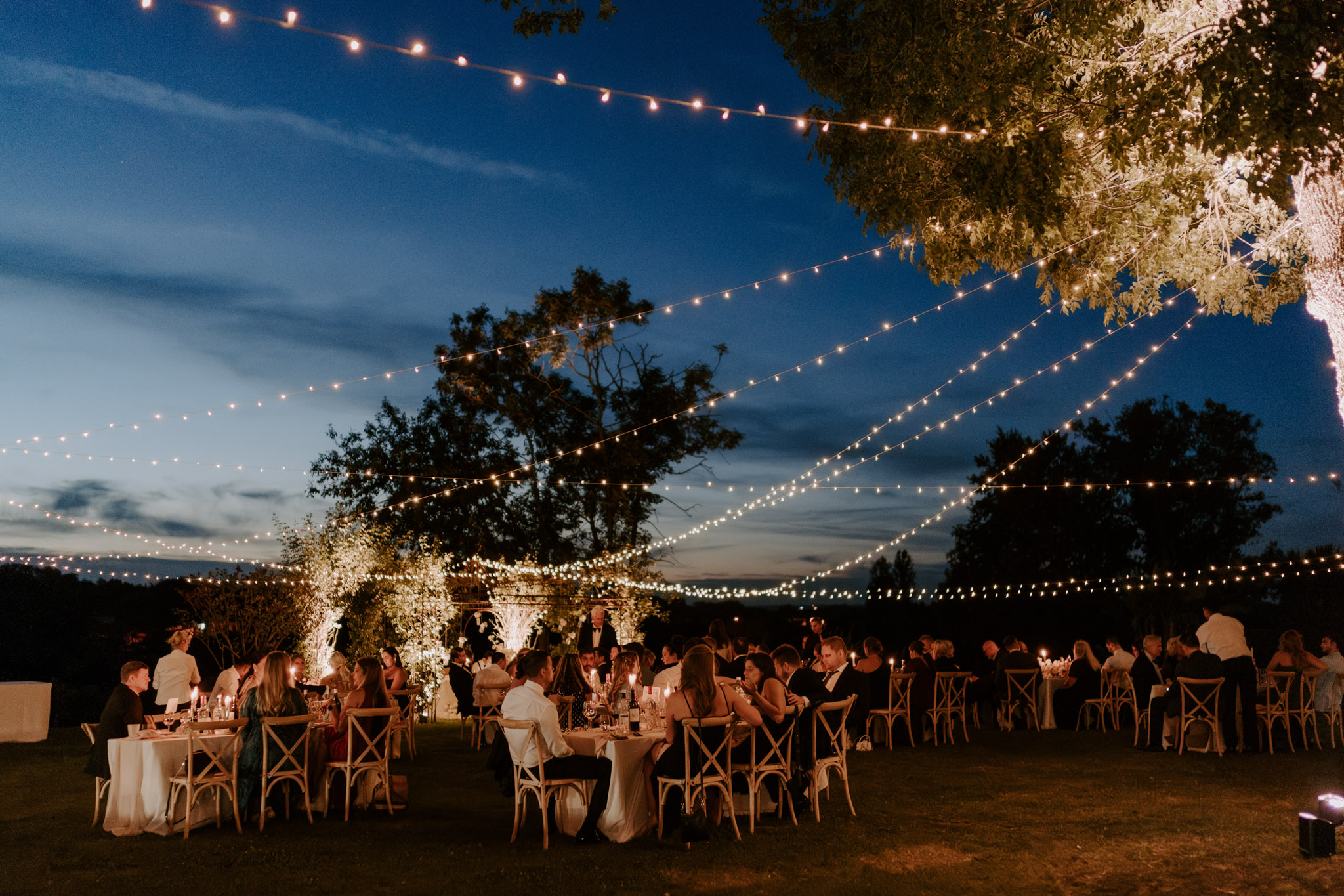 Evening reception with 70 guests at round tables under sweeping festoon lights against dusk blue sky