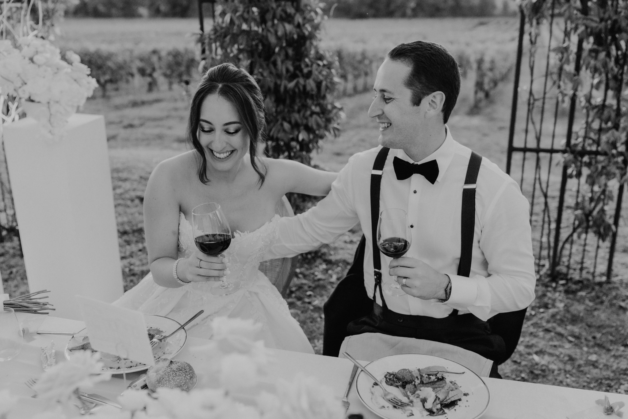 Black and white photo of bride and groom laughing with wine glasses at sweetheart table during vineyard reception