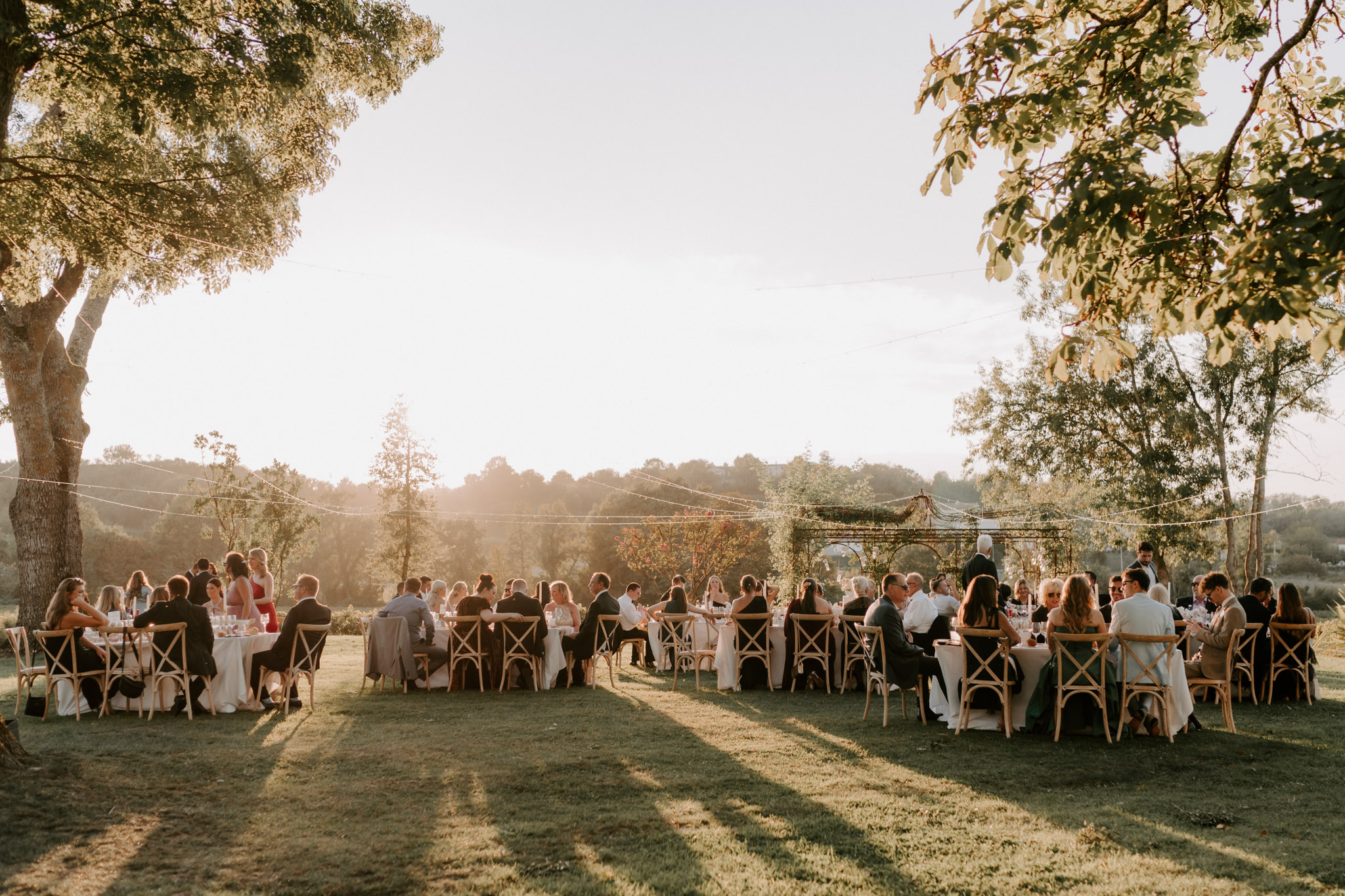 Sixty guests at round tables with cross-back chairs under festoon lights on lawn at golden hour