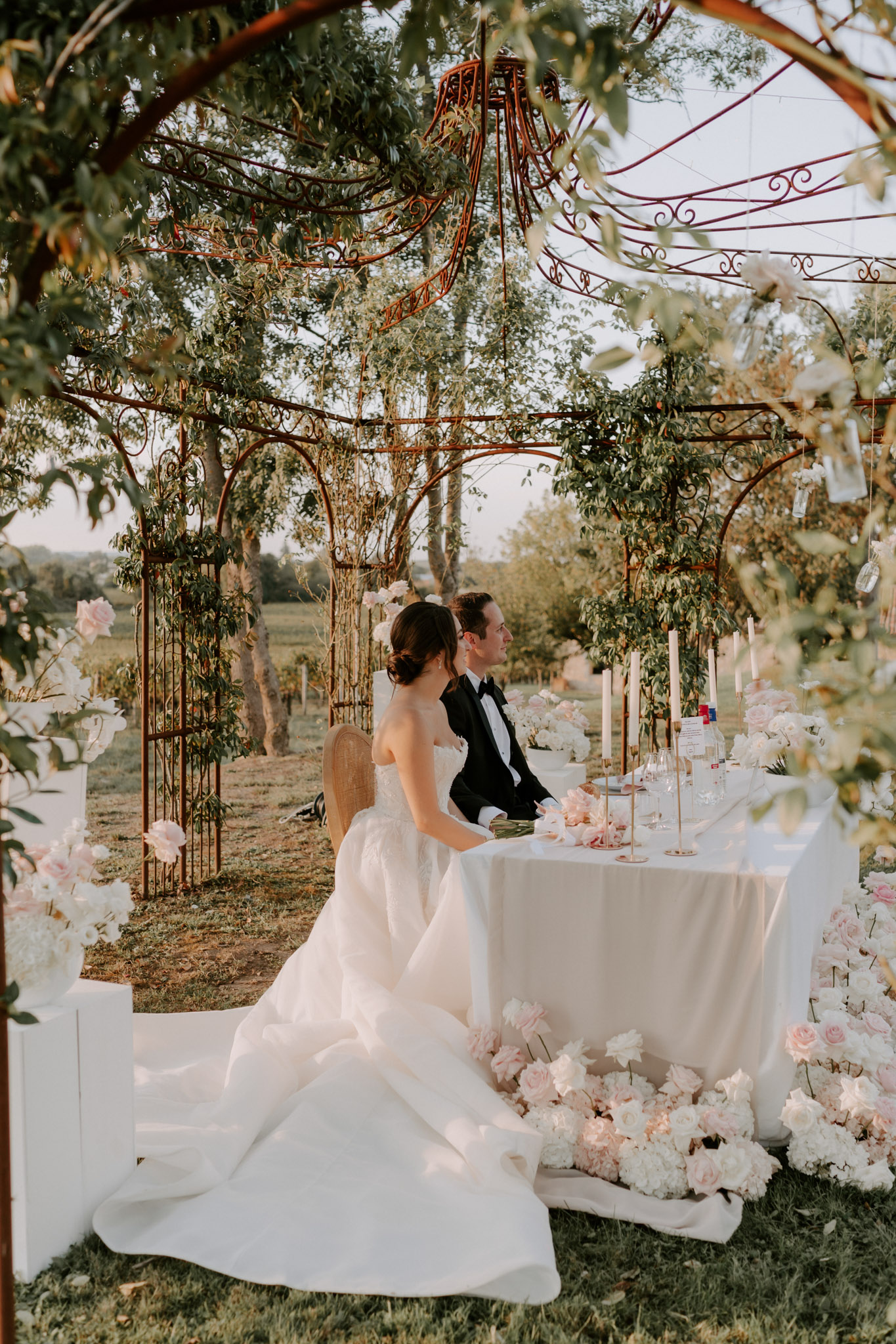 The bride and groom are seated together at a sweetheart table during an outdoor garden reception, likely at a vineyard or estate property. The bride wears a strapless ball gown with a full skirt and long train, and the groom is dressed in a black tuxedo with a bow tie. They are positioned beneath an ornate wrought-iron pergola structure with scrollwork detailing, draped with trailing greenery. The sweetheart table is covered in a white linen and styled with tall ivory taper candles in gold holders, crystal glassware, and scattered blush pink rose petals, while large arrangements of white hydrangeas, blush pink roses, and peonies are clustered on the ground around and beneath the table. Additional blush and white floral arrangements frame the left side of the frame near a decorative iron gate. The image is a medium-wide portrait shot taken during golden hour, giving the scene a warm amber tone, and is framed through foreground foliage for depth.