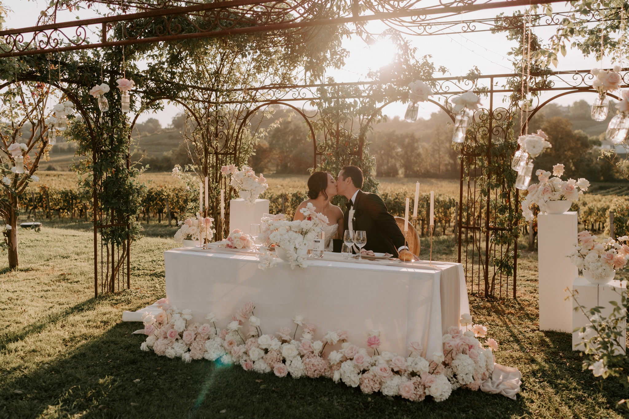The bride and groom share a kiss while seated at their sweetheart table during an outdoor reception at a vineyard, with rows of vines stretching across the background in warm golden hour light. The table is dressed in a white linen and bordered at the base by a dense floor-level arrangement of blush pink roses and white peonies, with additional tall taper candles and low floral centerpieces in the same blush and white palette on the tabletop. The couple sits beneath an ornate wrought iron pergola entwined with foliage, from which glass bud vases and clusters of white and blush blooms are hung at intervals. Two white tiered wedding cakes are displayed on white pedestals flanking the table, and the overall decor style is classic-romantic with a soft blush and white color palette.