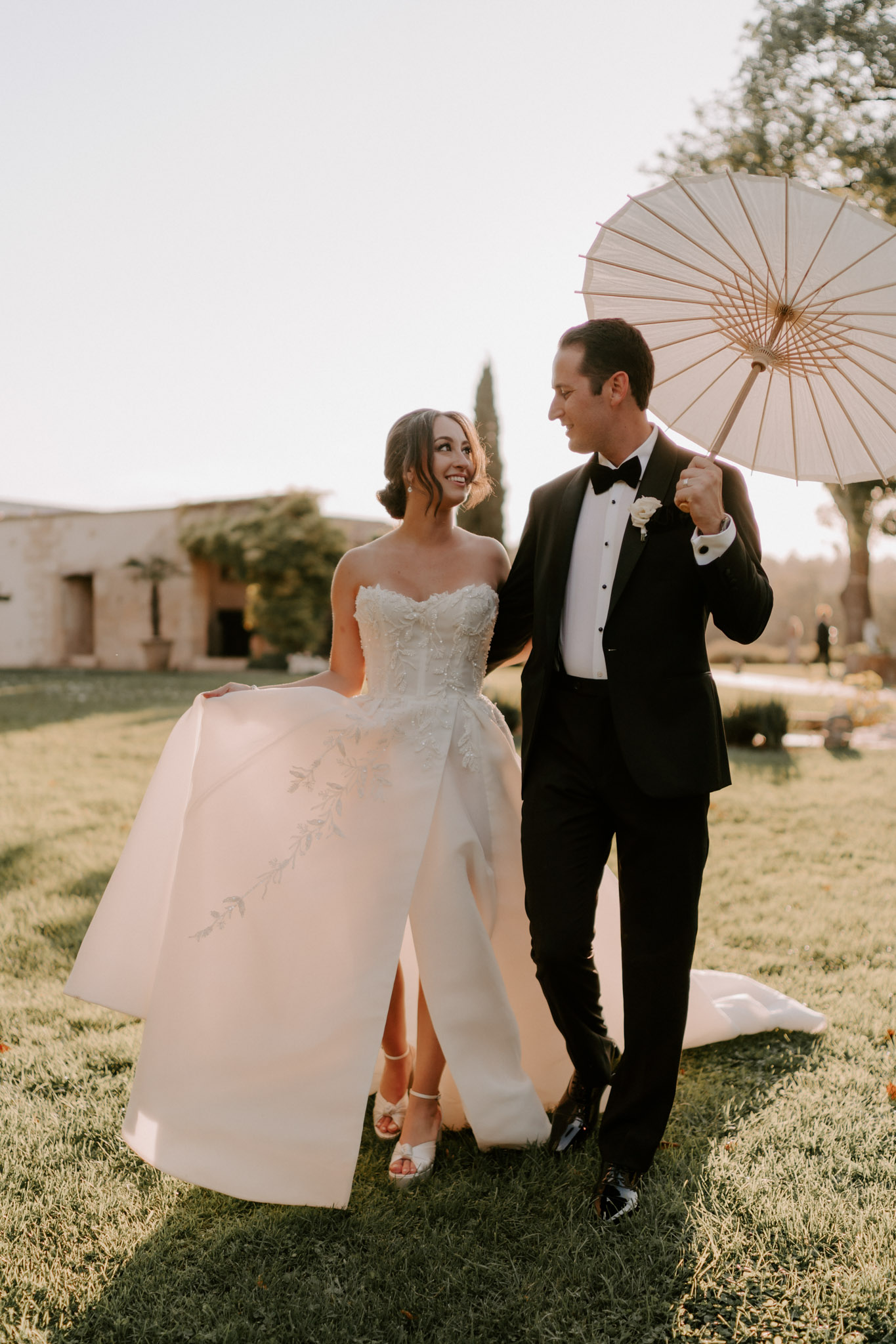 A couple portrait taken outdoors on a manicured lawn during golden hour, with a low stone building and cypress trees visible in the background. The bride wears a strapless ivory ball gown with a heavily embroidered lace bodice and a structured, dramatic overskirt featuring delicate floral embroidery on the skirt panels, paired with strappy silver heeled sandals and an updo hairstyle. The groom wears a black tuxedo with a black bow tie, white dress shirt, and a white rose boutonnière, and holds an ivory paper parasol over the couple. Both are looking at each other and smiling, with the bride lifting one side of her skirt. The composition is a full-length portrait shot with warm natural backlighting.