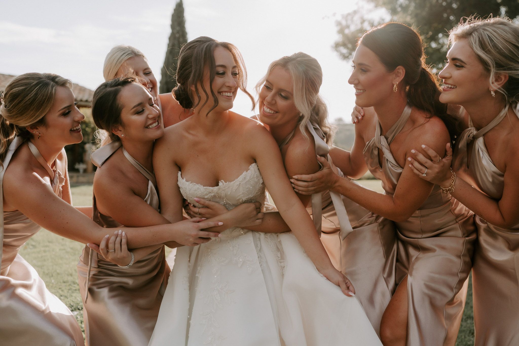 Bride in strapless lace ballgown surrounded by six bridesmaids in champagne and blush satin dresses laughing together