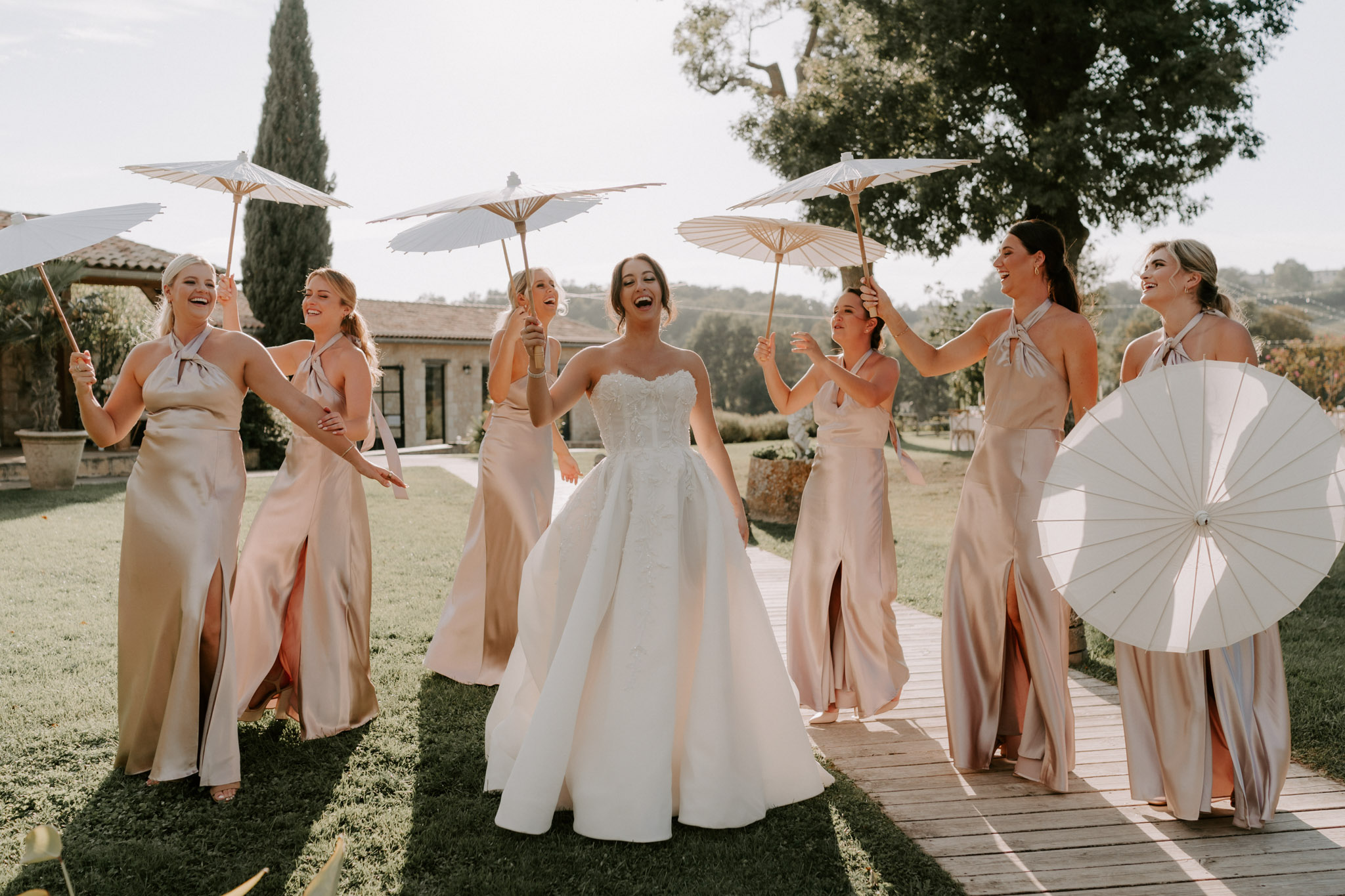 Bride and six bridesmaids in champagne satin dresses laughing with white parasols on Provencal estate deck