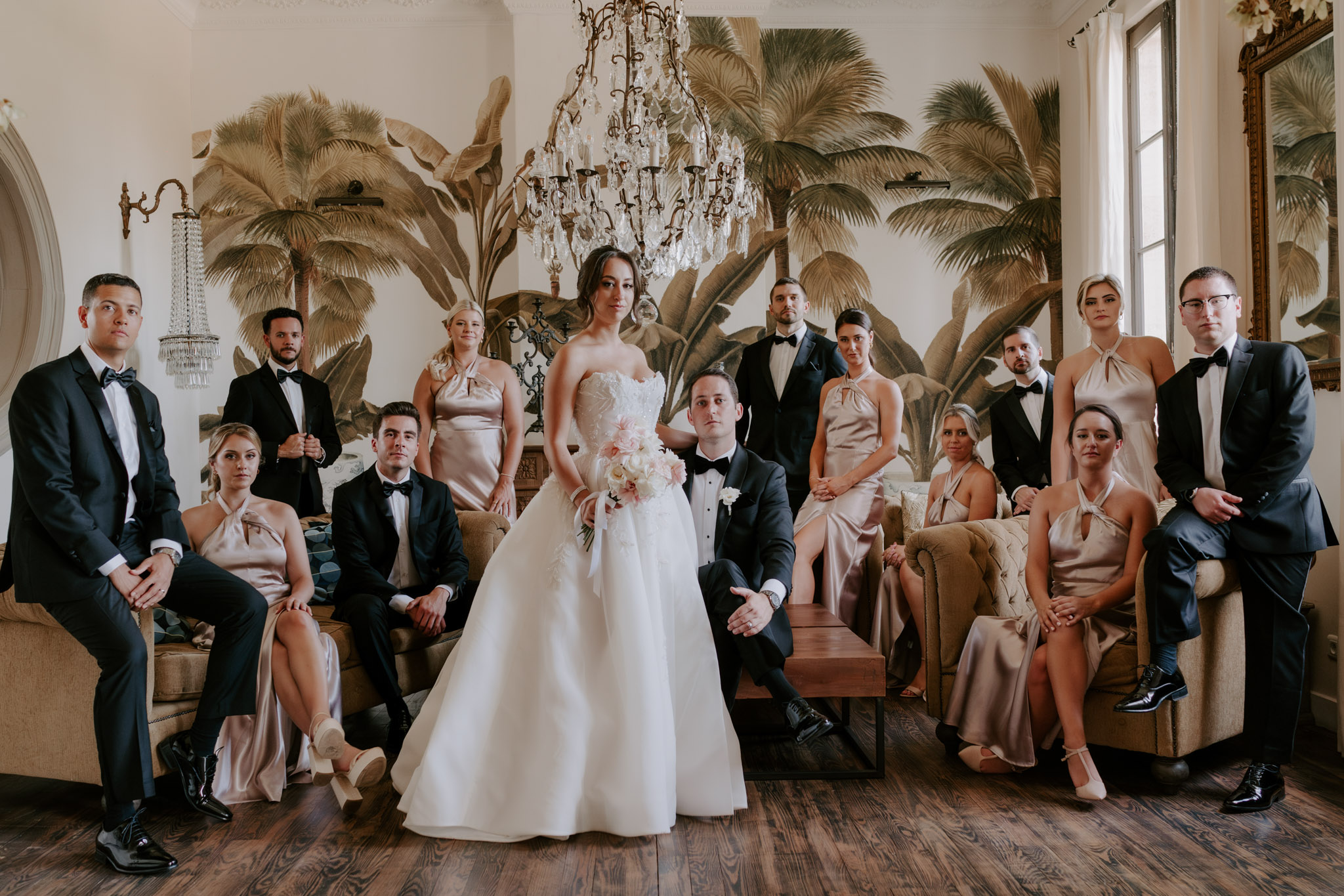 Full bridal party on velvet sofas in palm-mural salon with bridesmaids in champagne-gold satin dresses