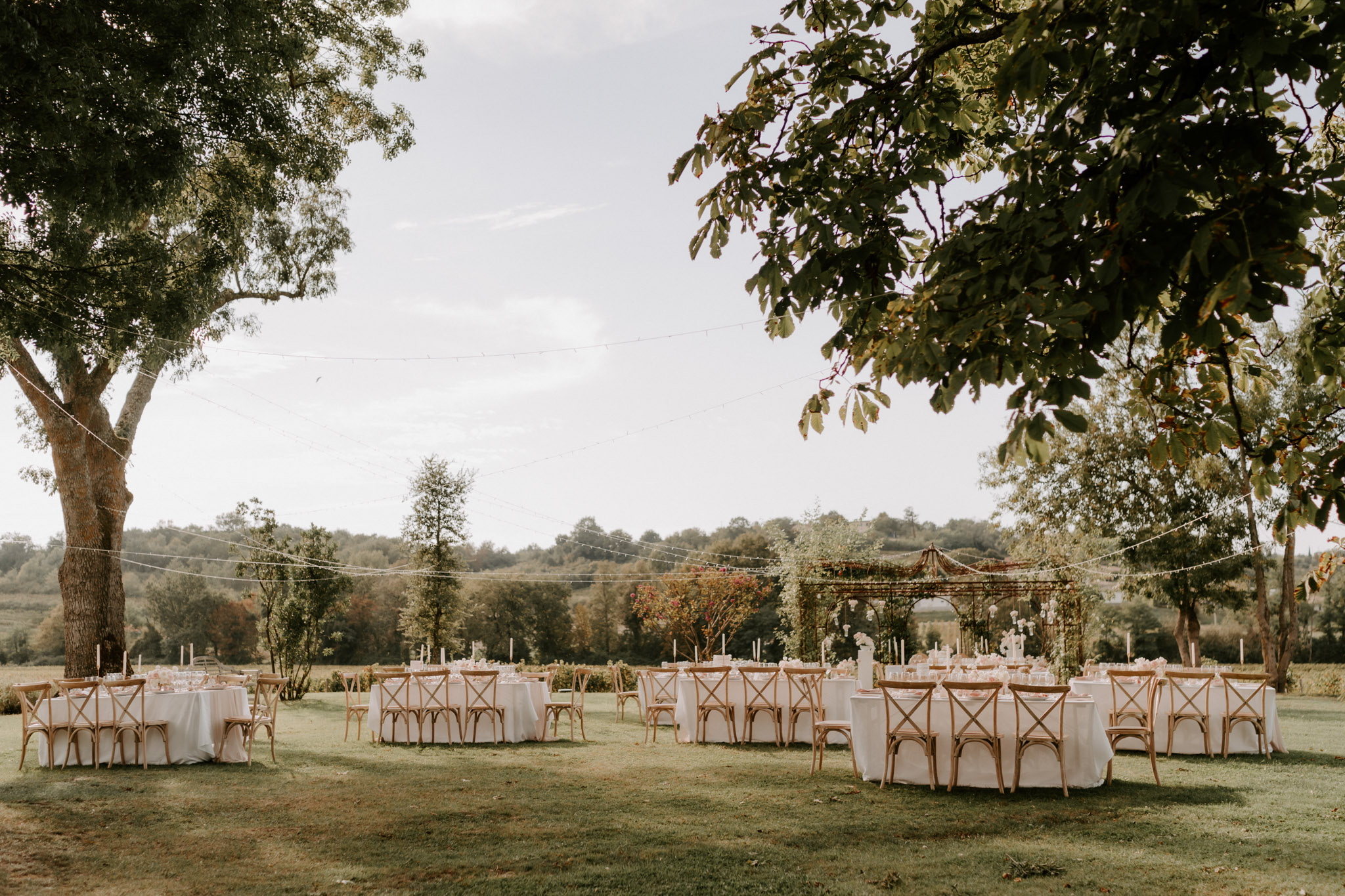 An outdoor wedding reception setup on a wide lawn, photographed as a wide establishing shot with no guests present. Approximately six to eight round tables are dressed in white linen tablecloths and surrounded by natural wood cross-back chairs, arranged across the grass. Each table is set with tall white taper candles, glassware, and small floral centerpieces in white and pale pink tones. Fairy lights are strung between the trees overhead, creating a canopy effect across the dining area. In the background, a wood and greenery pergola draped with flowering climbing plants serves as a focal decorative structure. The overall styling theme is rustic-romantic with a neutral and blush palette. Potential venue feature image.