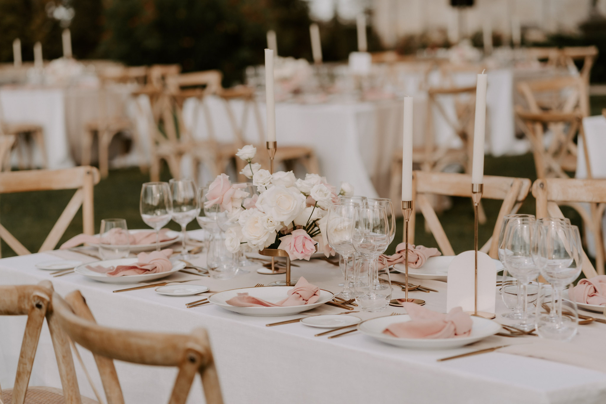 A close-up detail shot of an outdoor wedding reception table setup, with multiple additional tables visible and softly blurred in the background. The rectangular table is dressed in a white linen tablecloth and set with white ceramic plates, blush pink linen napkins, and gold flatware. Crystal wine glasses and water glasses are arranged at each place setting. The centerpiece is a low, compact arrangement of white and blush pink roses in a gold vessel, flanked by tall white taper candles in slim gold candlestick holders. Natural wood cross-back chairs surround the tables. The overall decor palette is white, blush pink, and gold, with a classic-modern styling approach.