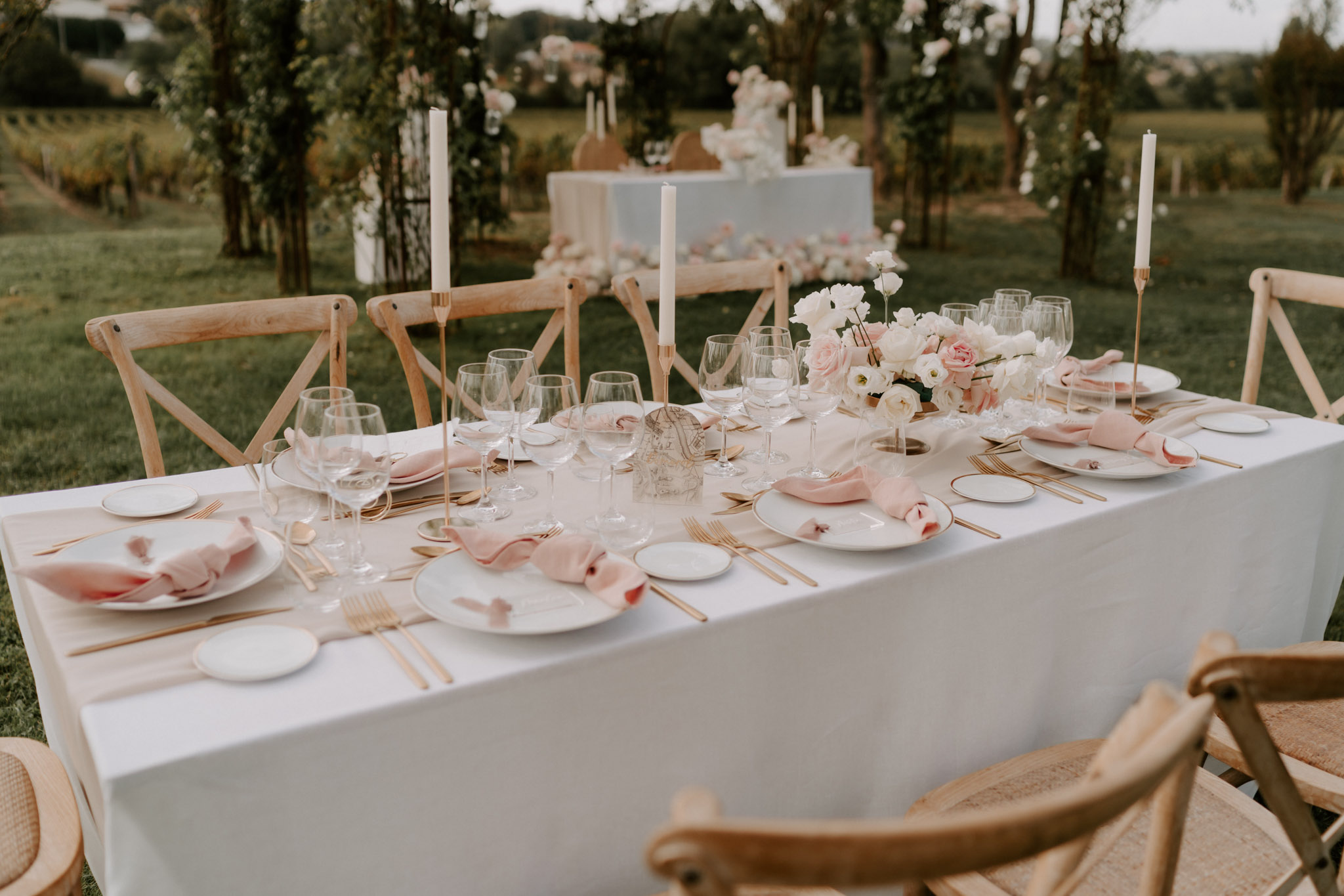 An outdoor wedding reception table setting photographed in a wide, slightly elevated angle, with a vineyard visible in the background. The long rectangular table is covered in a white linen tablecloth and set for approximately eight to ten guests, with white plates trimmed in gold, gold flatware, and blush pink linen napkins loosely folded on each place setting. Multiple clear wine glasses are arranged at each setting, and slender white taper candles in gold brass holders are spaced along the table. A low floral centerpiece of blush pink garden roses, white ranunculus, and white blooms runs along the middle of the table. Natural wood cross-back chairs surround the table. In the background, a separate white-clothed table displays what appears to be a tiered wedding cake decorated with white flowers, framed by foliage-wrapped structures. The overall decor palette is white, blush pink, and gold, consistent with a classic romantic outdoor reception style.