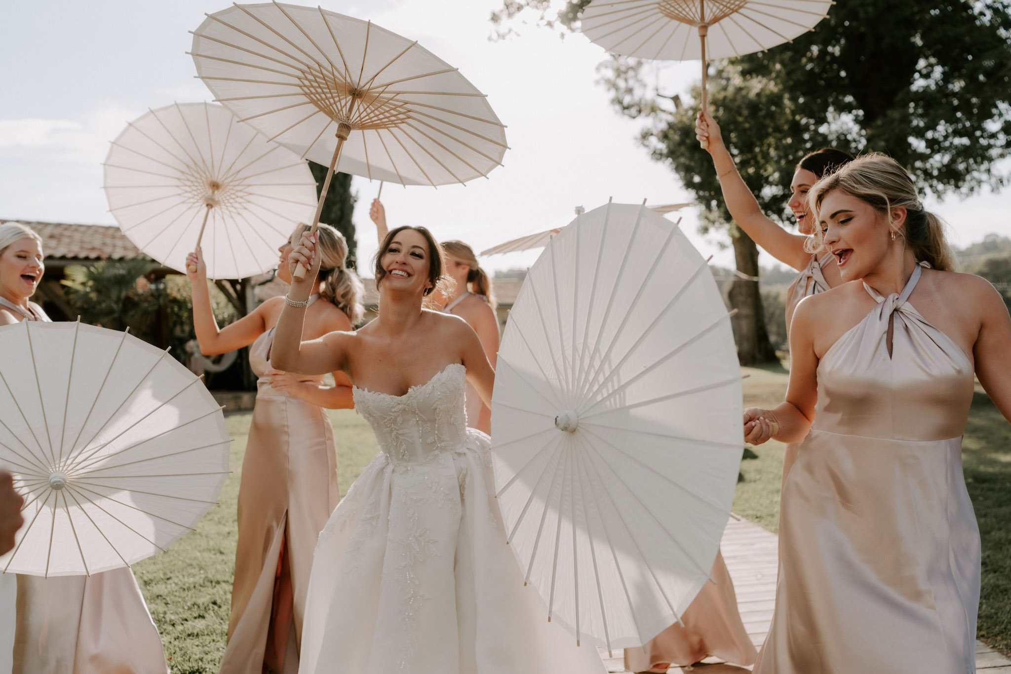 The bride and her bridal party of approximately five bridesmaids are walking and celebrating outdoors on the grounds of what appears to be a French countryside property with a terracotta-roofed building visible in the background. The bride wears a strapless, full-skirted white gown with floral appliqué and lace detailing, while the bridesmaids wear floor-length champagne/blush satin halter-neck dresses with front slits in varying wrap and tie styles. Each person holds a white paper parasol with bamboo handles, creating a playful, coordinated styling element throughout the group. The shot is a candid mid-distance portrait capturing movement and laughter, taken in bright natural sunlight with a warm golden tone.