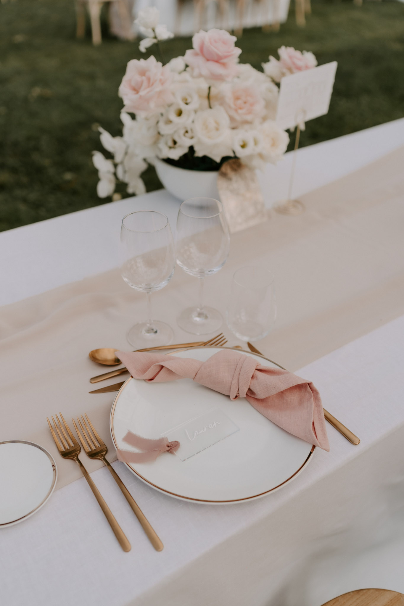 Close-up detail shot of an outdoor wedding reception table setting. The place setting features a white plate with a thin gold rim, a dusty rose linen napkin tied in a loose knot, a small white place card with cursive script reading 'Lauren', and gold flatware including two forks, a knife, and a spoon arranged on either side. Two clear wine glasses are positioned above the plate, and a beige table runner runs down the center of a white tablecloth. In the background, a white ceramic bowl holds a centerpiece of blush pink roses and white lisianthus blooms. The overall decor palette is white, blush pink, and gold, consistent with a modern classic styling approach.