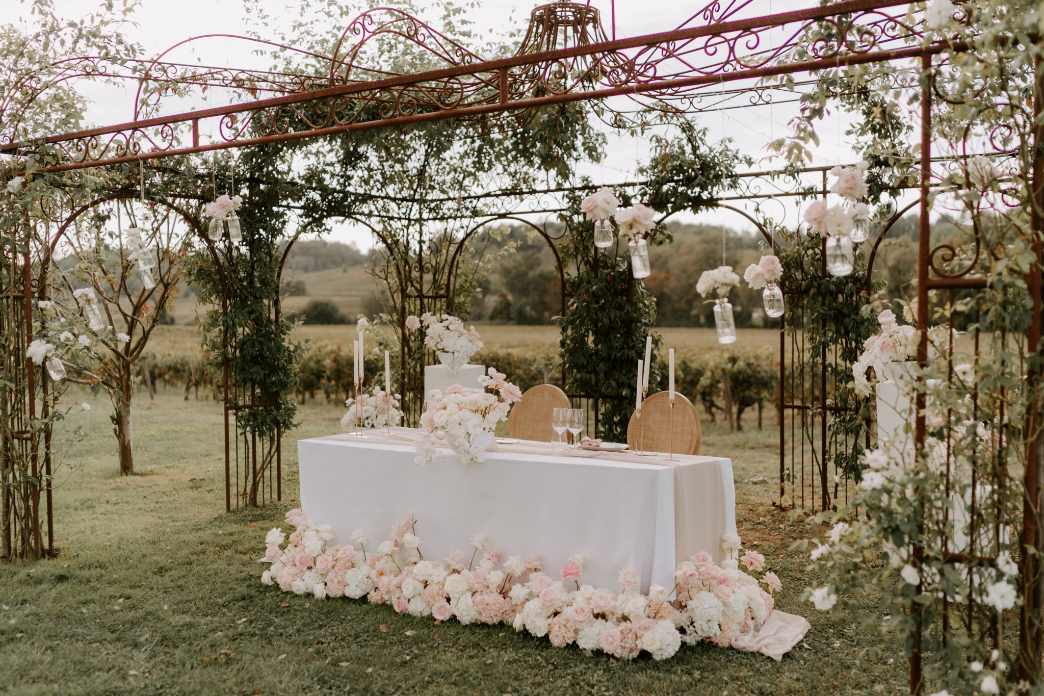 Sweetheart table under wrought-iron pergola with blush roses, ivory peonies, gold taper candles, and tiered cake overlooki...