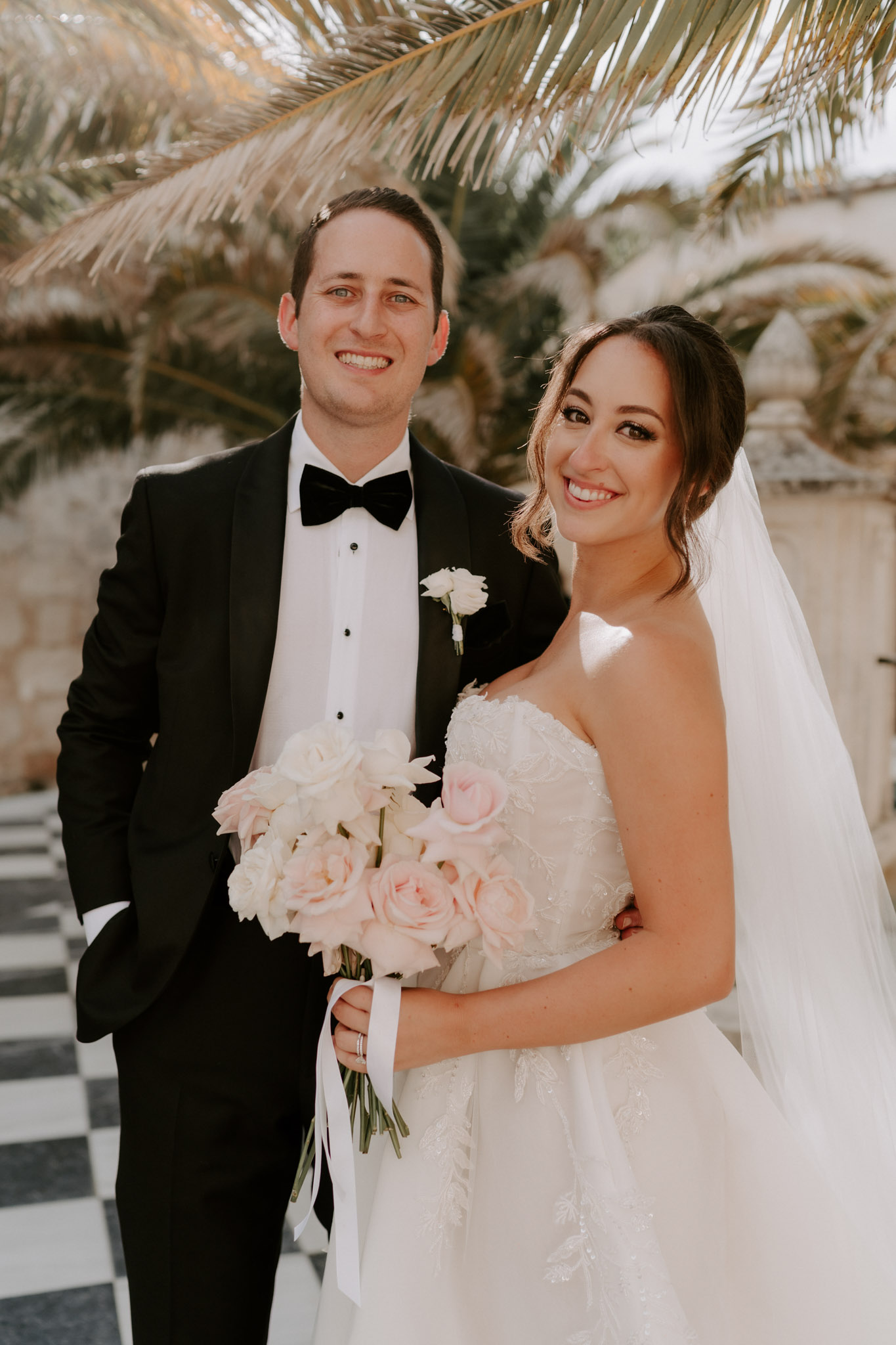 A couple portrait taken outdoors, with the bride and groom posed together and facing the camera. The groom wears a black tuxedo with a white dress shirt, black bow tie, and a white boutonniere. The bride wears a strapless, lace-embroidered ball gown in ivory with a cathedral-length veil and an updo hairstyle. She holds a rounded bouquet of blush pink garden roses and white flowers tied with a blush satin ribbon. The setting features a black-and-white checkered tile floor and a stone architectural backdrop with palm fronds visible overhead, suggesting a formal outdoor courtyard. The overall styling is classic and formal, with a black-and-white and blush pink color palette.