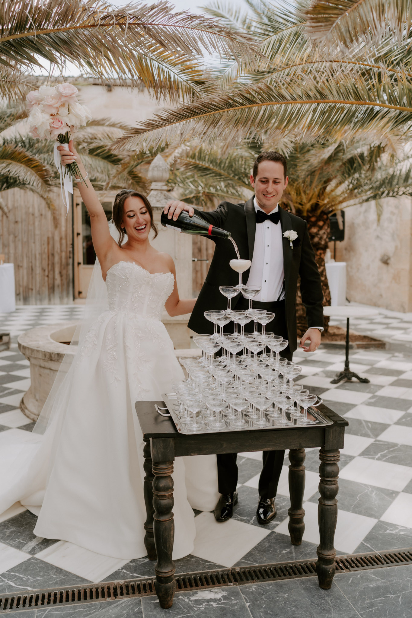 The bride and groom are pouring a champagne tower during what appears to be a cocktail hour or reception, set in an outdoor courtyard with a black-and-white checkered marble floor. The groom, wearing a black tuxedo with a black bow tie and a white boutonniere, pours champagne from a bottle into the top coupe glass of a pyramid tower arranged on a dark wooden table. The bride, in a strapless ivory ballgown with lace bodice detail, raises her blush and white bouquet — composed of blush roses and white blooms with trailing white ribbon — triumphantly in the air while laughing. The setting features stone architecture, a large decorative stone basin in the background, and palm trees overhead, giving a warm Mediterranean feel to the venue. The overall styling is classic and formal, with the black tuxedo, monochrome flooring, and champagne tower contributing to a refined black-and-white palette. This is a full-length portrait shot of the couple.