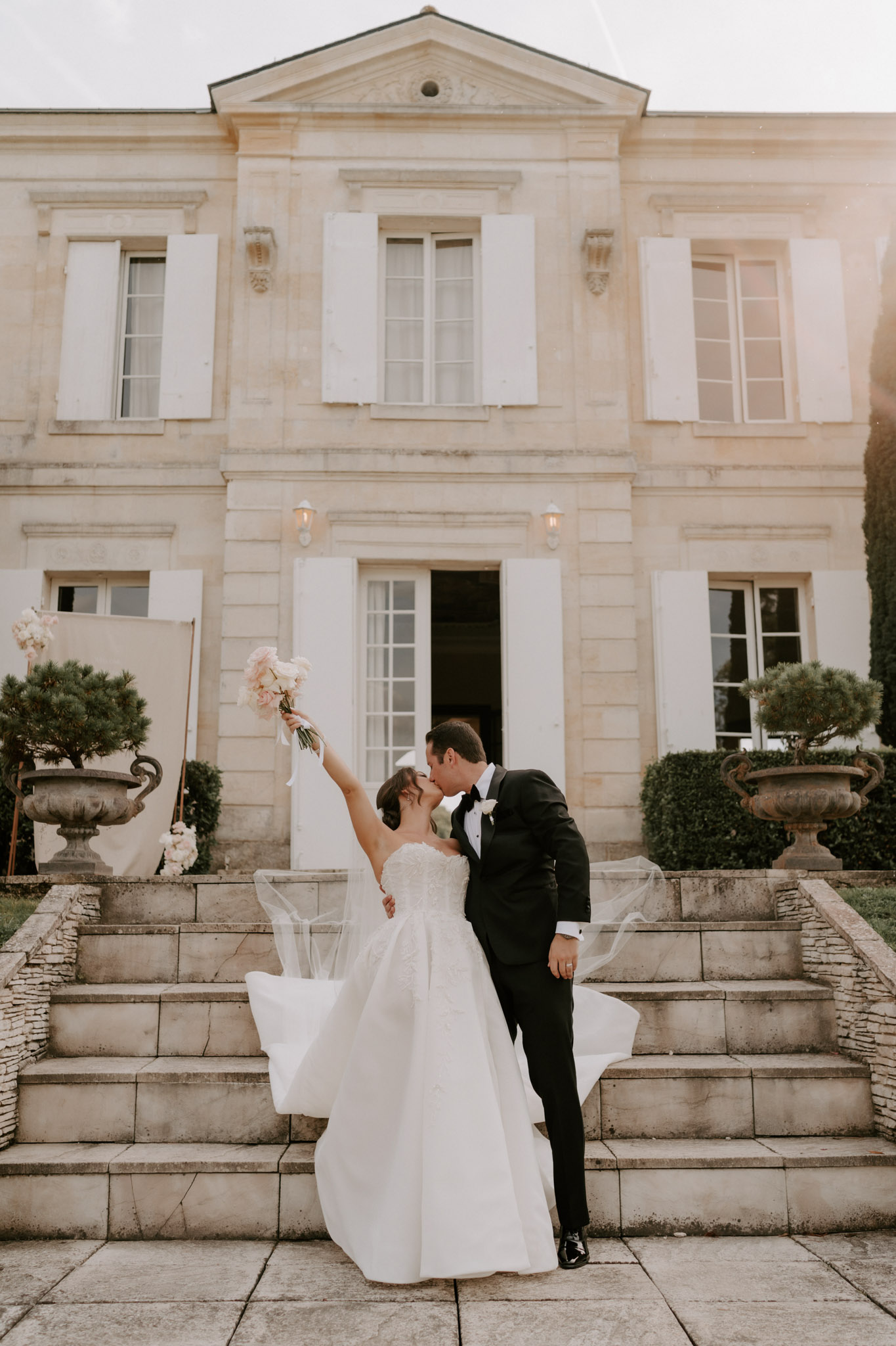 Bride raising peony bouquet overhead kissing groom on chateau steps in golden light