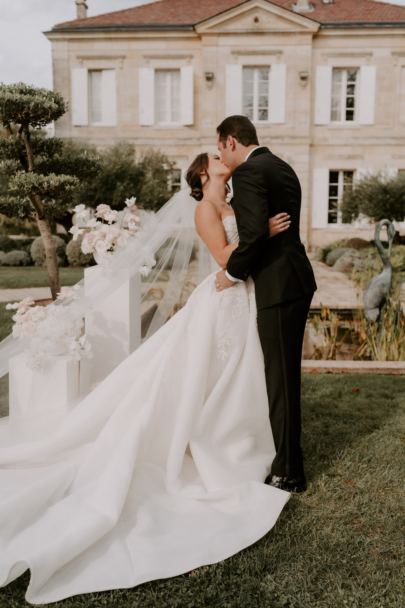 A couple shares a kiss during outdoor portraits on the grounds of a French château, identifiable by its classic limestone façade, red-tiled roof, and white shuttered windows. The bride wears a strapless ivory ball gown with lace appliqué detailing on the bodice and skirt, a full cathedral-length train spread across the lawn, and a long flowing veil caught by a breeze. The groom is dressed in a classic black tuxedo with a white dress shirt. To the left of the couple, white geometric rectangular plinths of varying heights hold arrangements of blush pink and white florals — including what appear to be ranunculus and garden roses — serving as a ceremony backdrop structure. The composition is a full-length portrait shot with the château facade centered in the soft-focus background, and bronze bird sculptures visible in the formal garden to the right. Potential venue feature image.