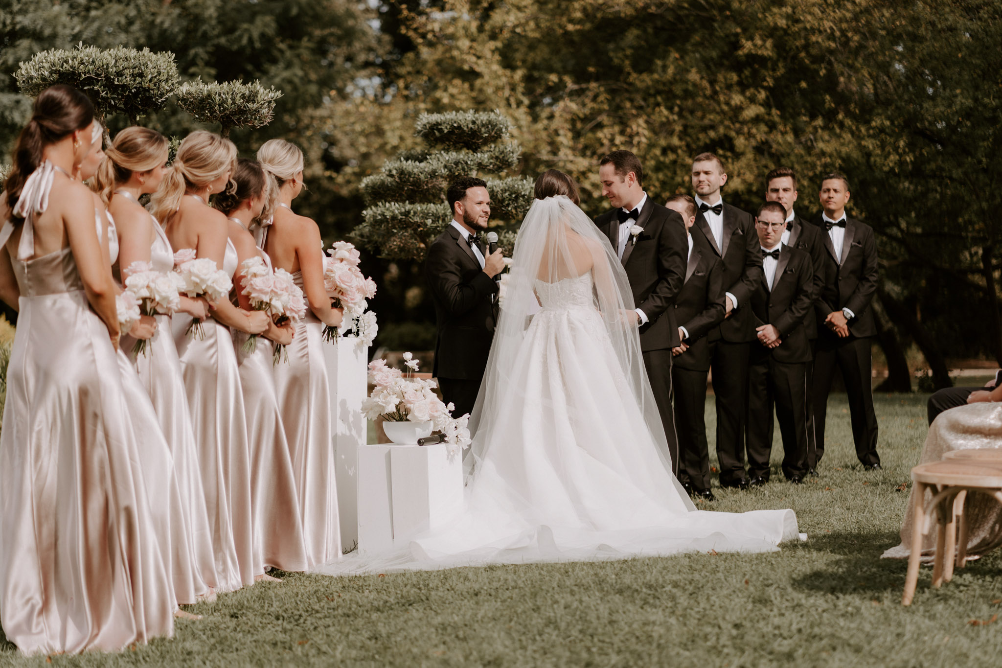 Couple at altar with five bridesmaids in blush satin and five groomsmen before white geometric plinths