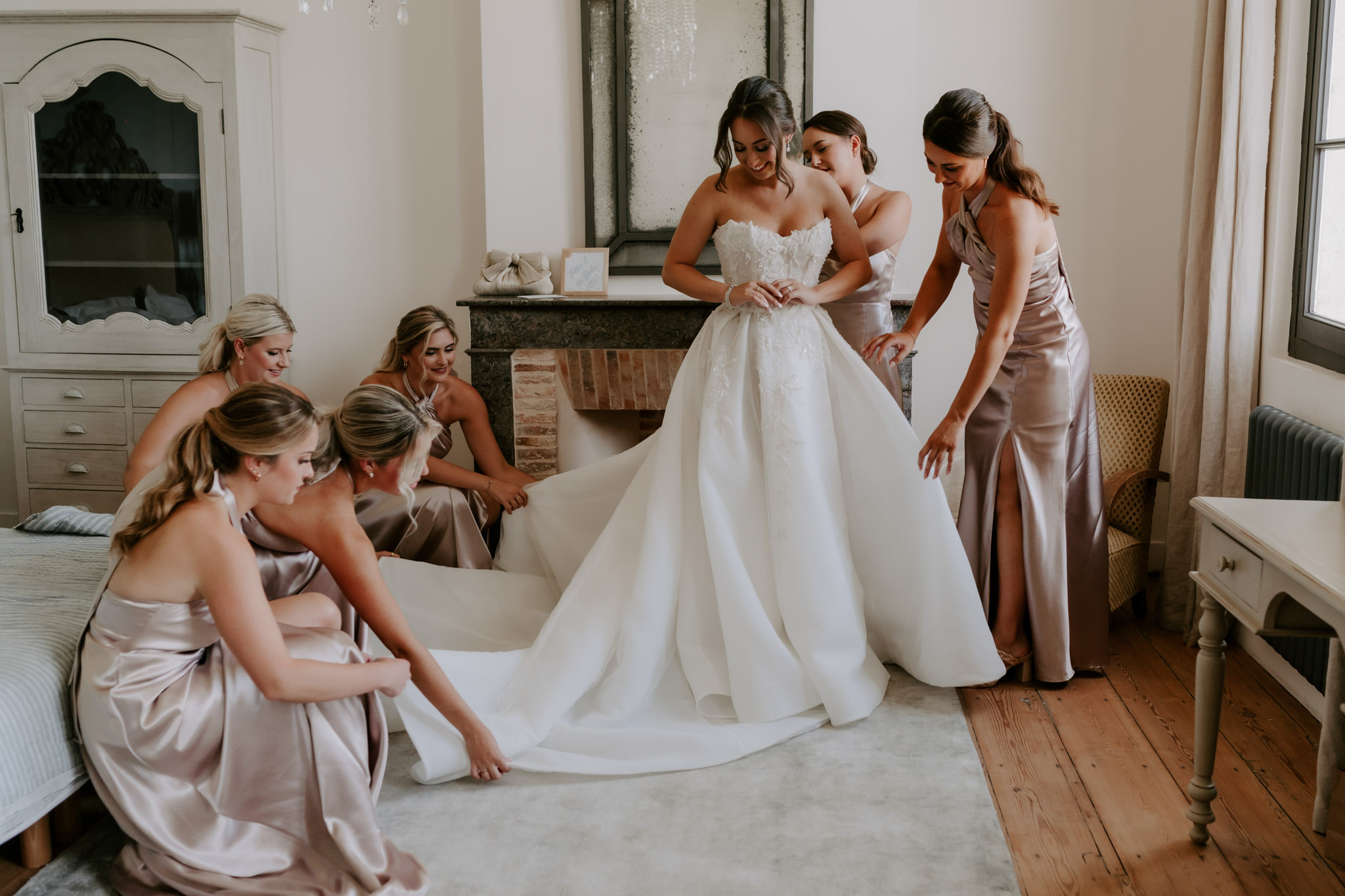 Five bridesmaids in dusty mauve dresses arranging bride's feathered ballgown train in chateau bedroom