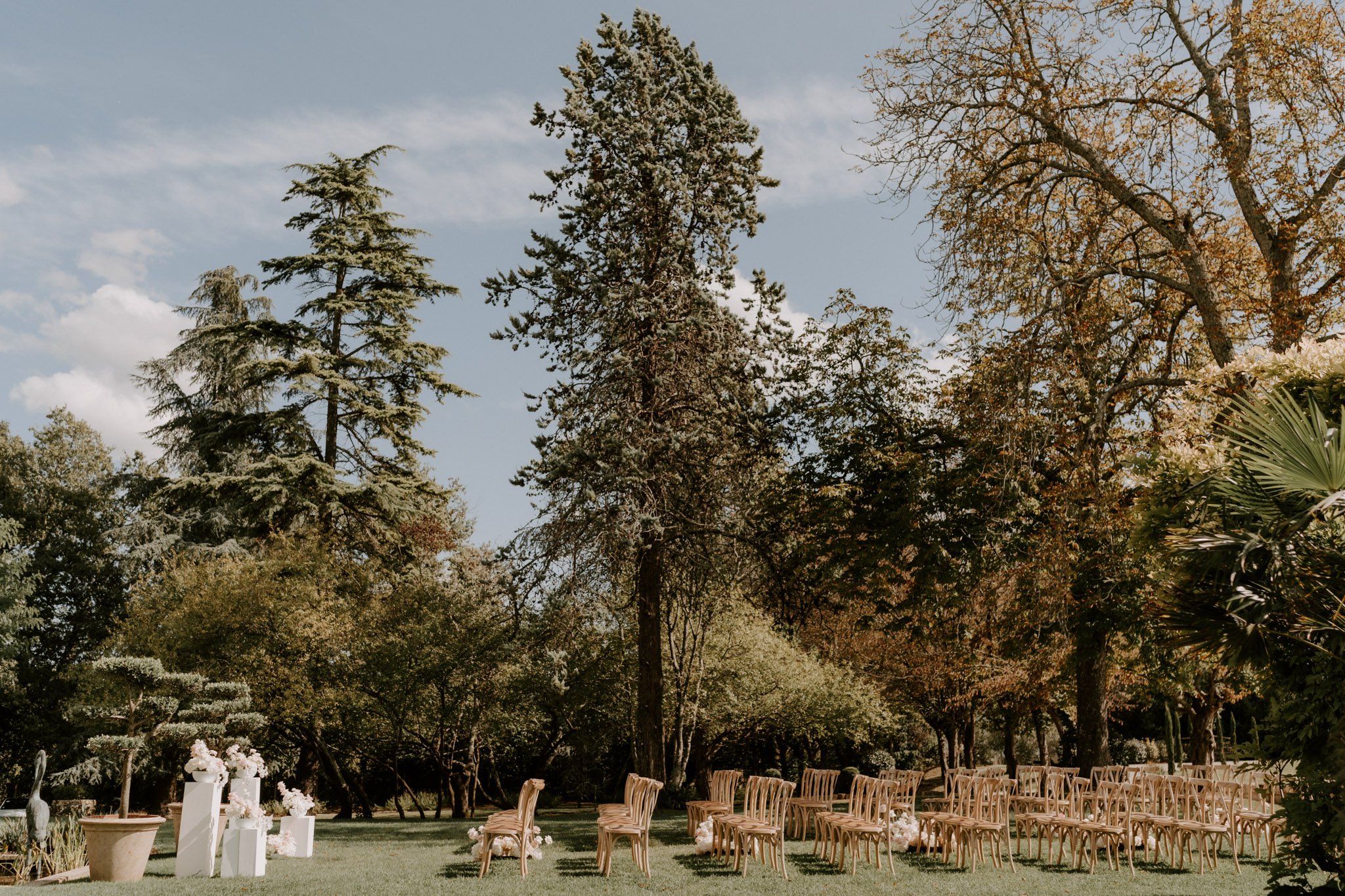 Outdoor ceremony setup with cross-back wooden chairs and white pedestals displaying blush and ivory floral arrangements